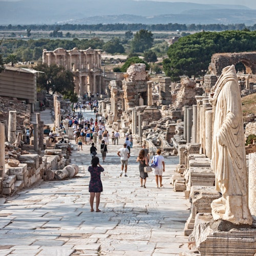 Tourists explore ancient ruins featuring columns, statues, and a large stone pathway, with a mountainous backdrop.