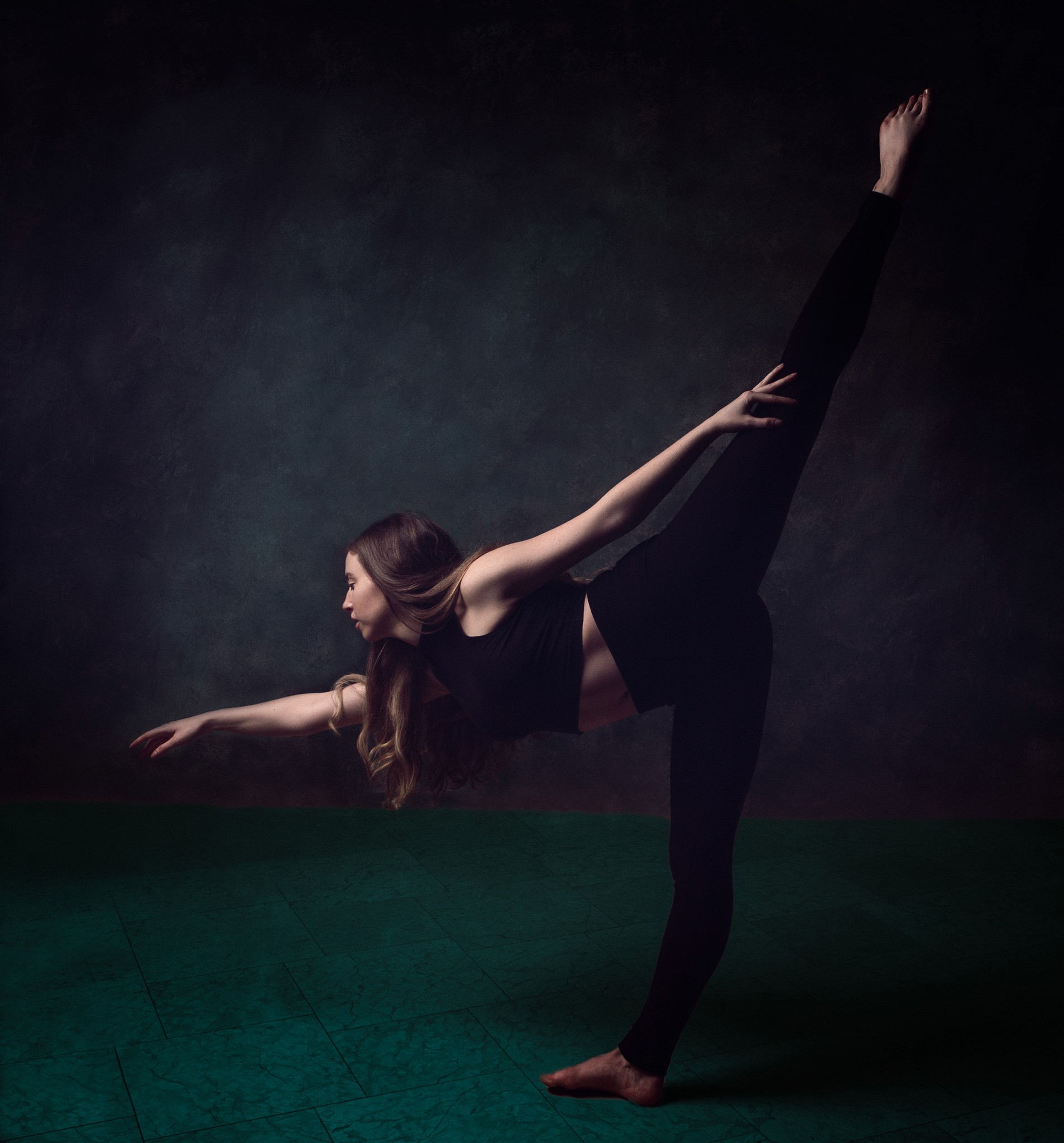 Studio portrait of a ballet dancer balancing on one leg with the other extended behind her, captured in dramatic side lighting against a dark textured backdrop.