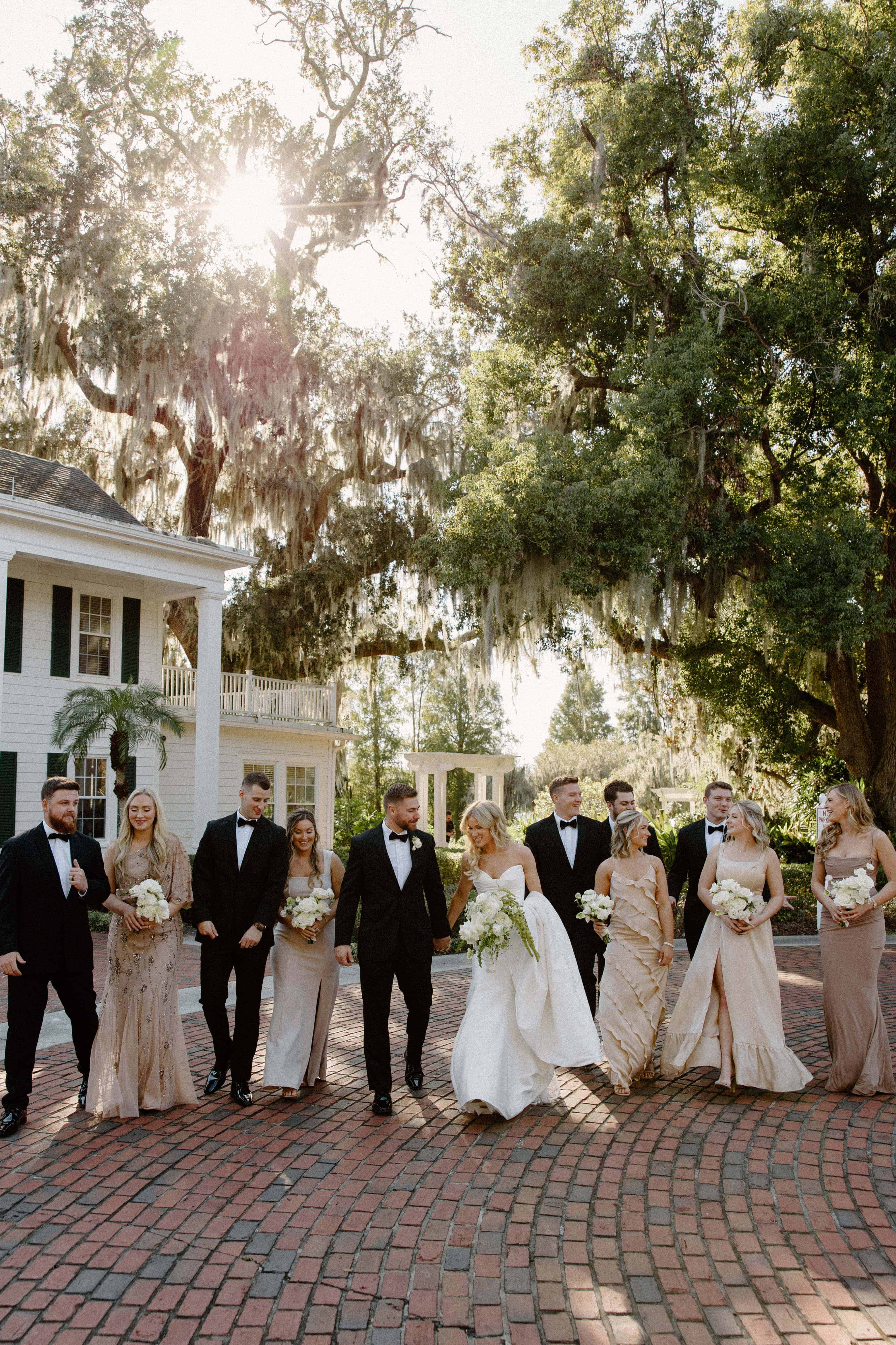  A photo of a wedding party standing on a brick walkway in front of a white house. The men are in tuxedos and the women are in long dresses.
