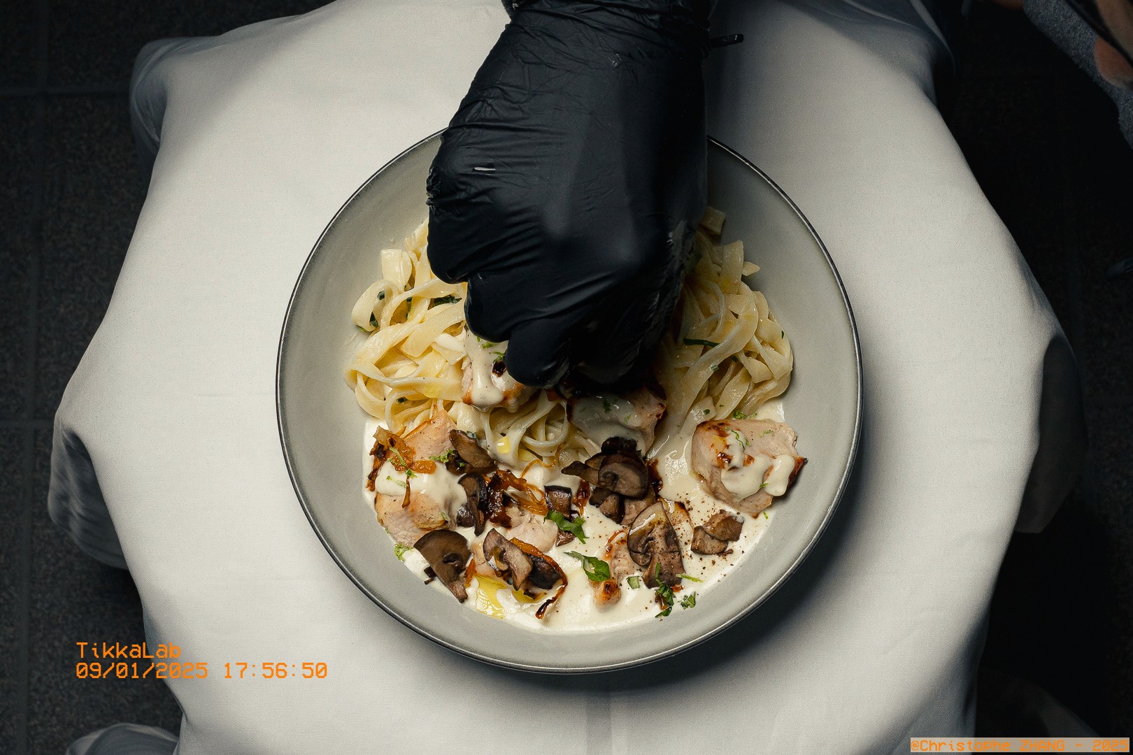 Assiette de tagliatelles crémeuses avec poulet et champignons, dressée par une main gantée de noir.