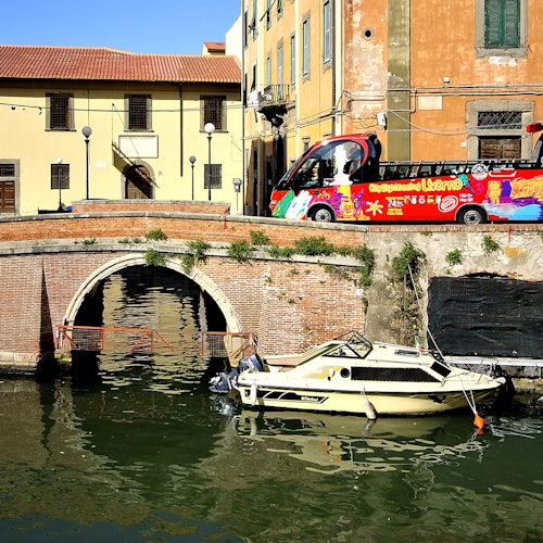 小船停靠在砖砌拱桥旁的运河边,背景是色彩缤纷的旅游巴士和建筑物。