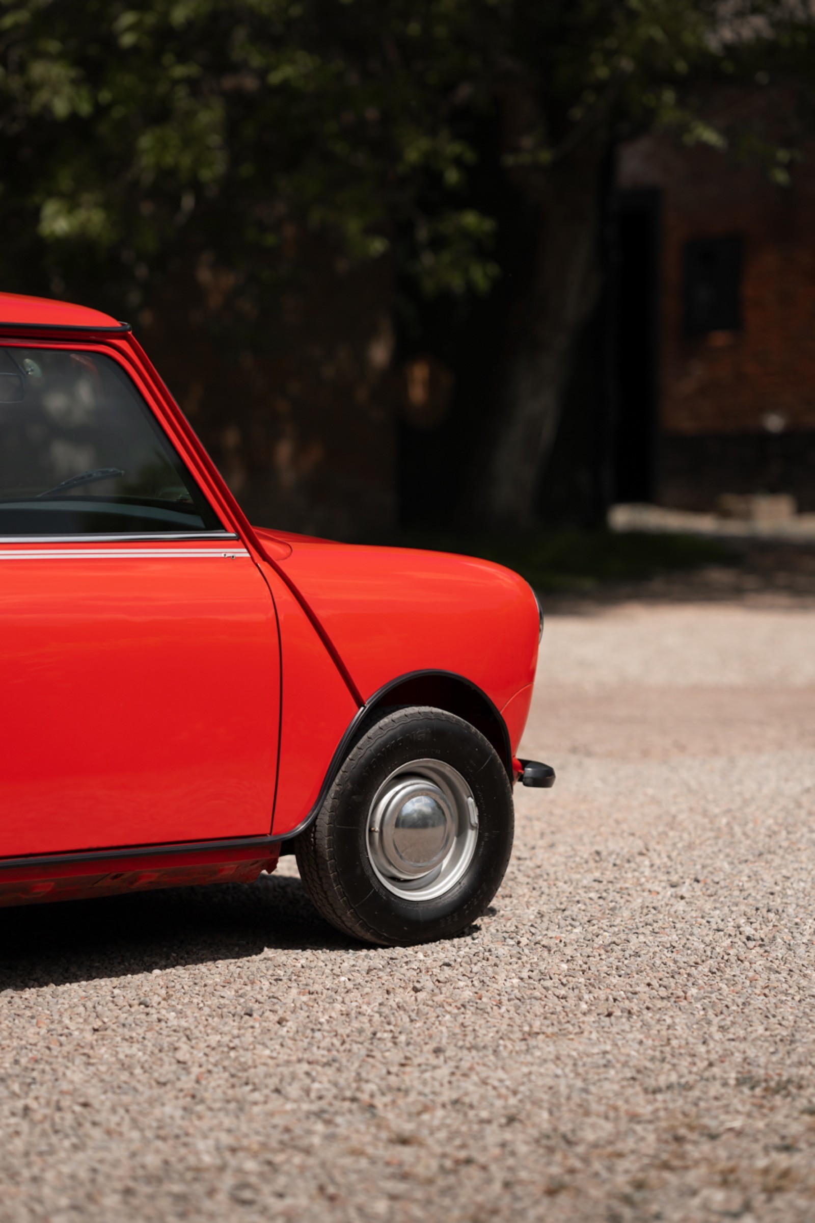 A close-up of a red vintage car's front end parked on a gravel surface, with blurred background greenery.