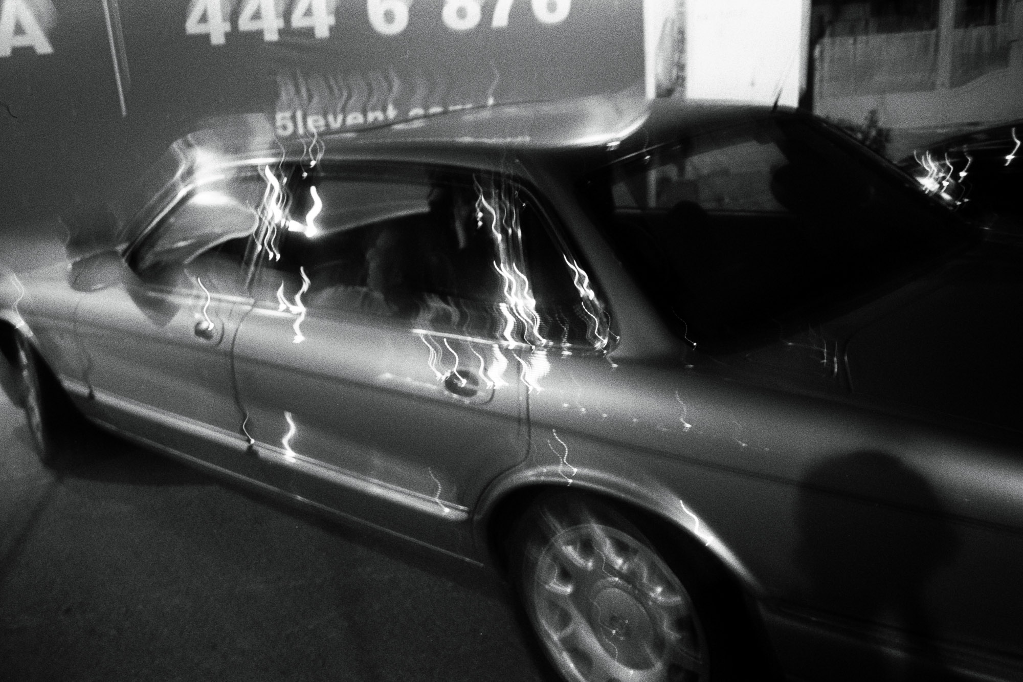 A classic sedan is parked under dim lighting, casting a sleek silhouette with reflections on its polished surface, surrounded by blurred motion and illuminated signs, creating a dynamic urban night scene.
