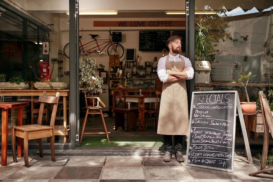 Bearded barista in apron stands outside coffee shop entrance with "We Love Coffee" sign and specials board.