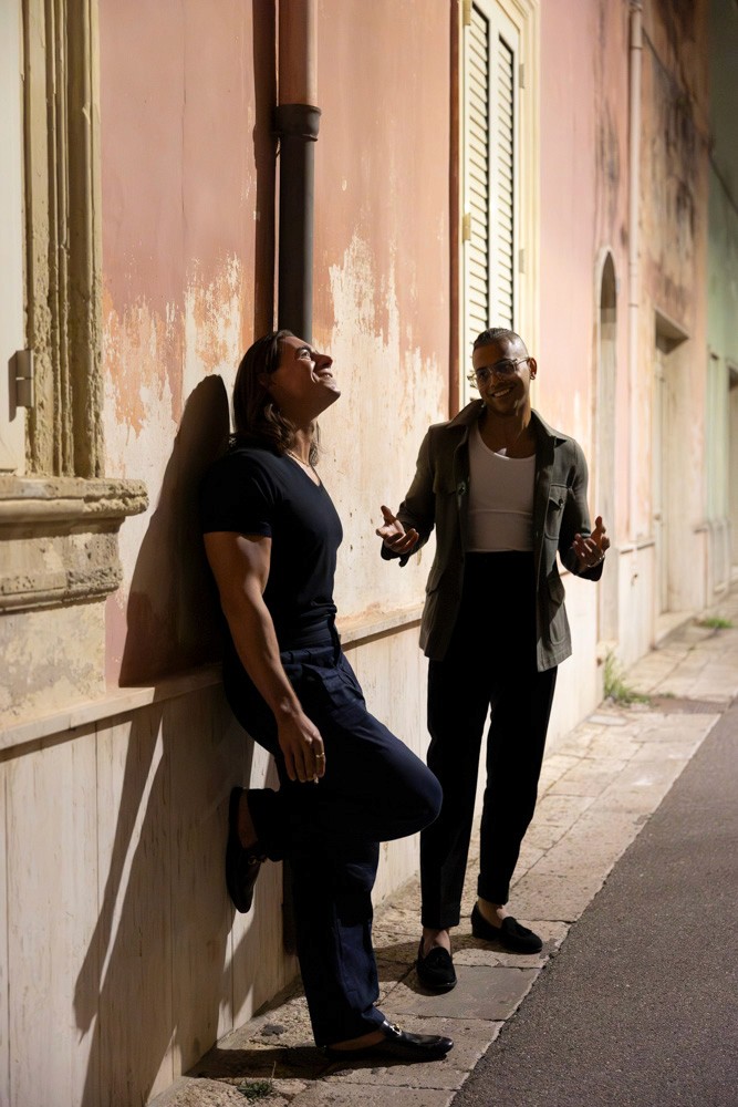 Two men dressed in elegant evening attire sharing a relaxed conversation along a softly lit street, embodying refined confidence and modern sophistication.
