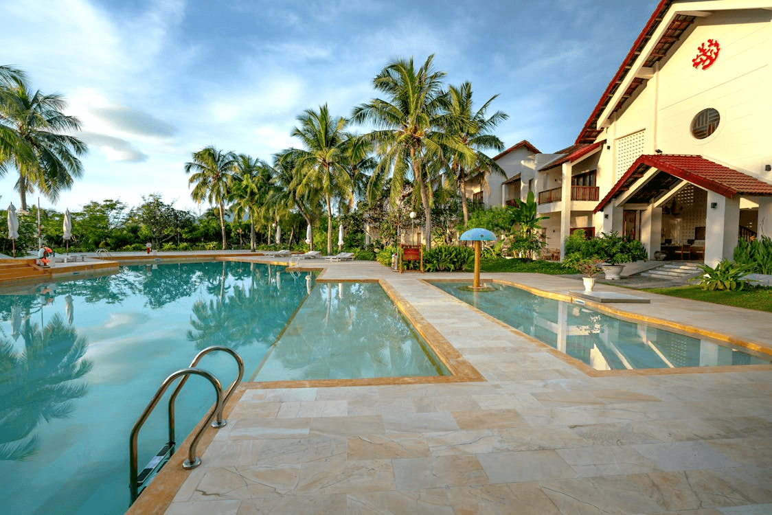 Tropical resort with yellow buildings, palm trees, umbrellas, and large lagoon-style pool surrounded by landscaping.
