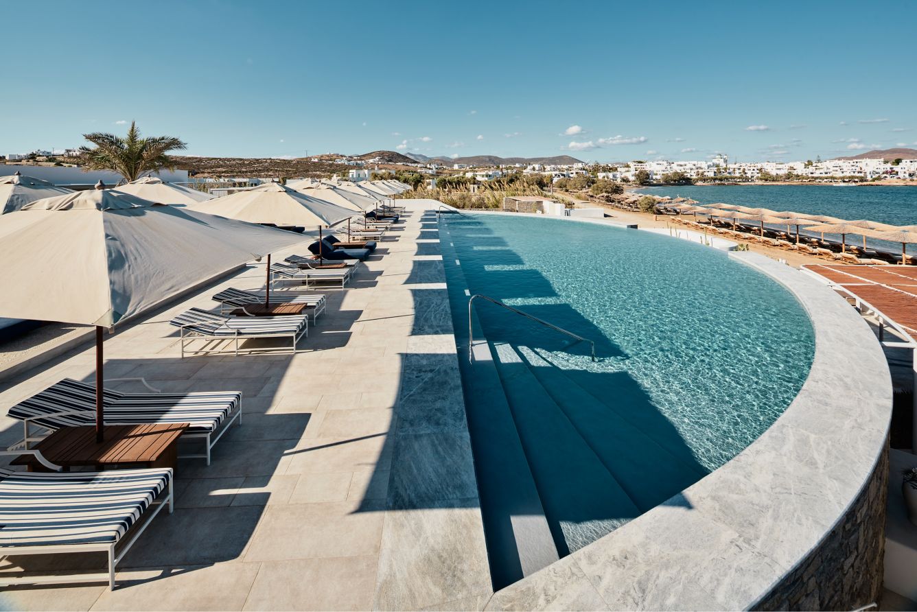 Large, curved infinity pool with a long row of striped sun loungers and umbrellas right on a bay.