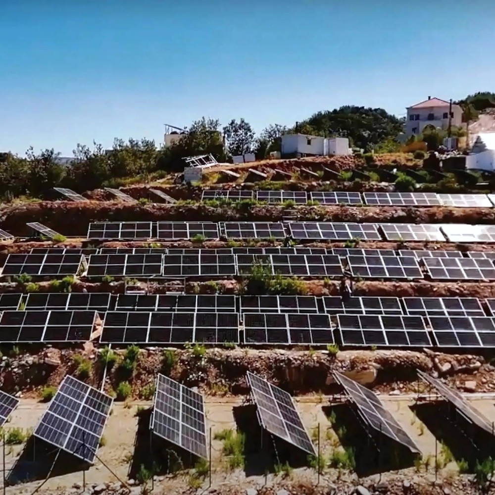 A hillside solar farm with numerous solar panels arranged in neat rows, surrounded by greenery and under a clear blue sky, providing sustainable energy in a scenic landscape.