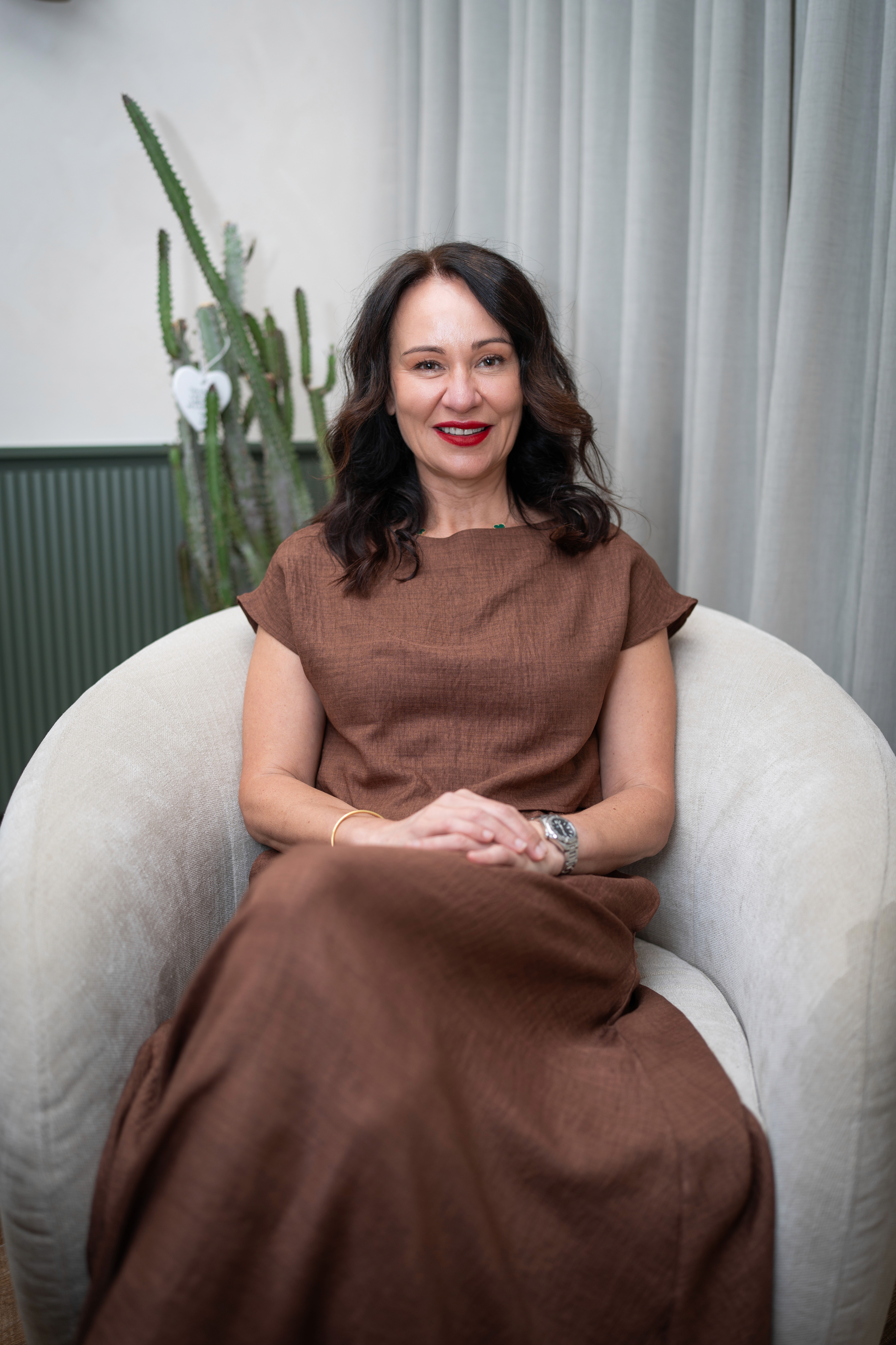 Dr. Bonnie Hawthorne in a brown dress sits relaxed on a white chair, surrounded by greenery and soft curtains in the background.