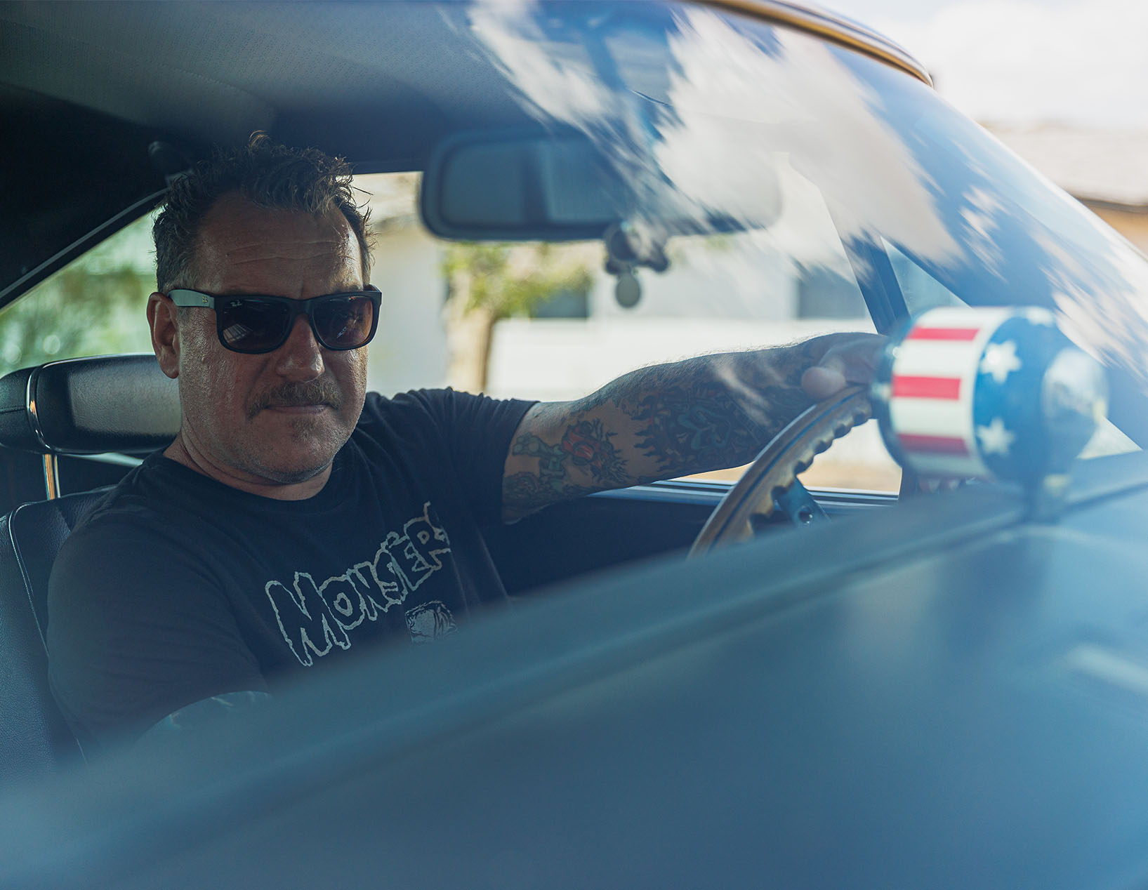Dirty Donny GIllies wearing sunglasses and a “Monsters” T-shirt sitting behind the wheel of a classic car, seen through the windshield with reflections of blue sky and clouds.