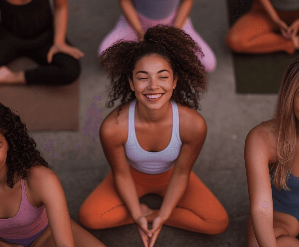 Smiling woman in a yoga pose, wearing a white tank top and orange pants, surrounded by others on mats. The scene conveys calm and happiness.