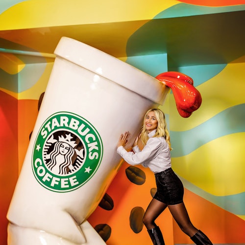 Woman posing with oversized Starbucks coffee cup amidst colorful, abstract background. Cup spilling red liquid.