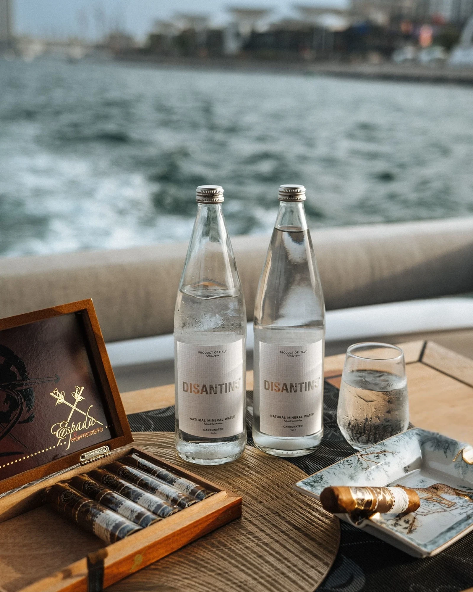 Two bottles of Disantino mineral water on a wooden table beside an open cigar box, drinking glass, and cigar ashtray with water and boats visible in the background.