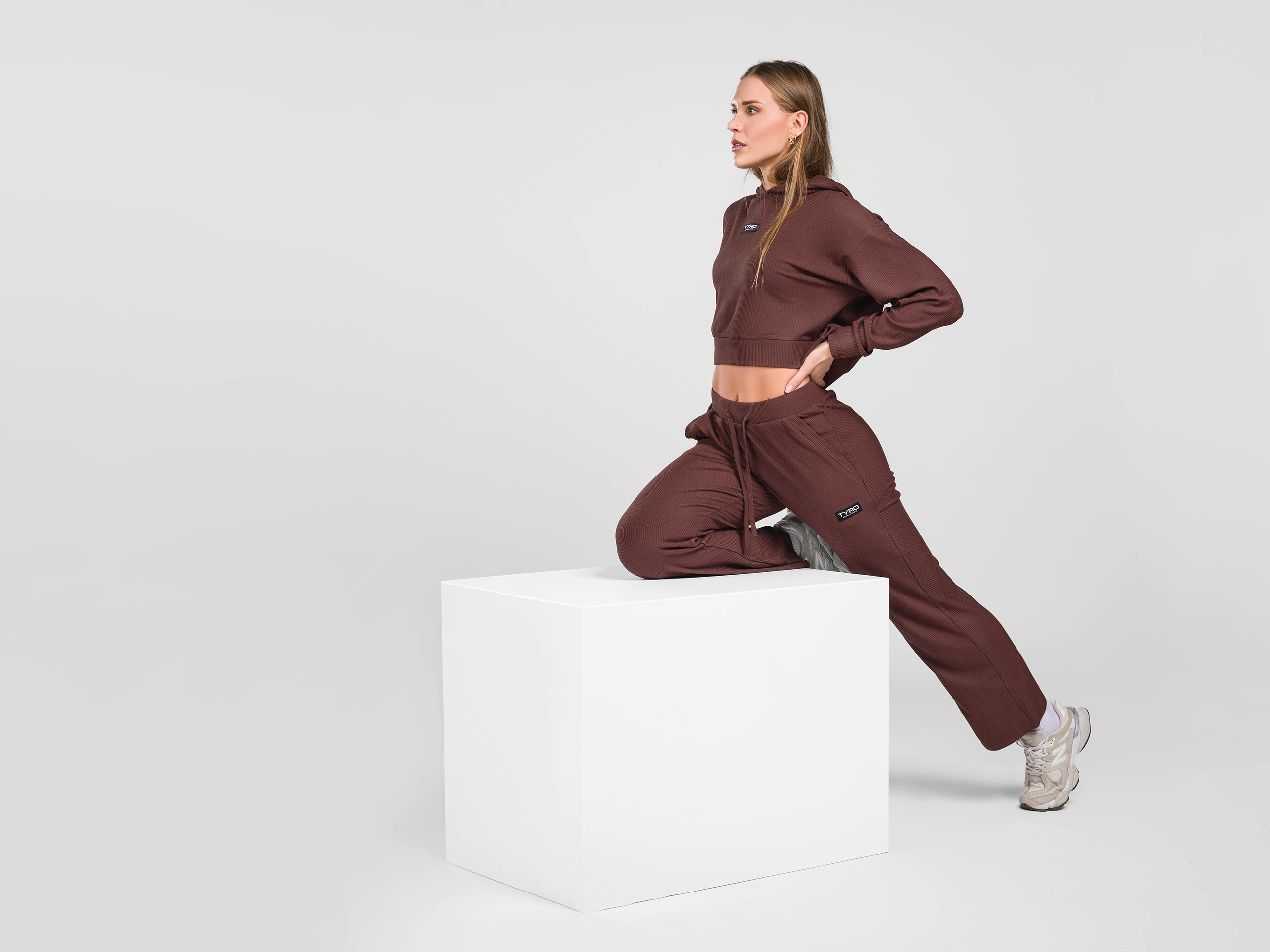 Woman in brown sportswear posing with one knee on a white cube in a studio.