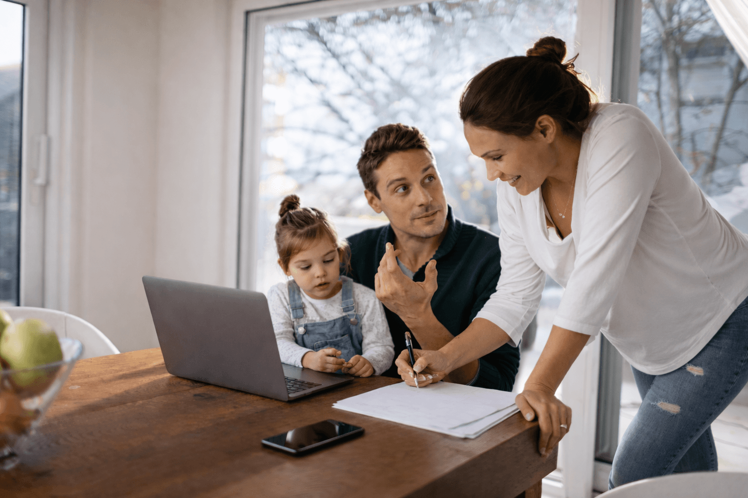 Family at a table with a laptop and papers, one parent signing a document while discussing it with the other.