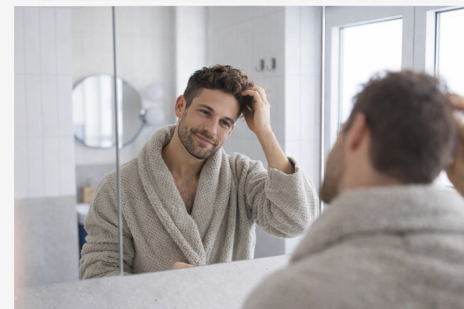 Man in a bathrobe looking at himself in a bathroom mirror and adjusting his hair.