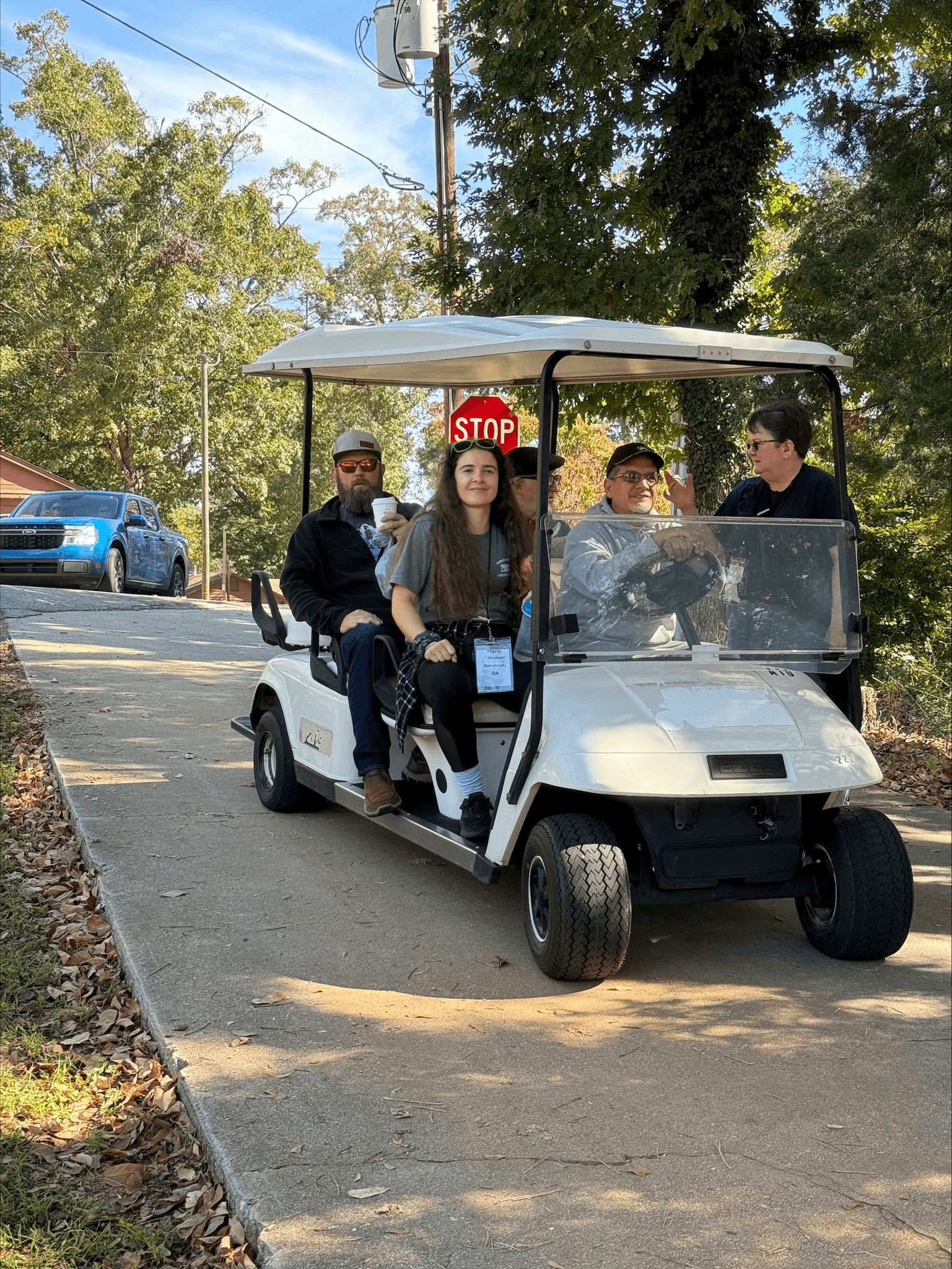 A group of people are riding in a white golf cart on a paved road under a blue sky.