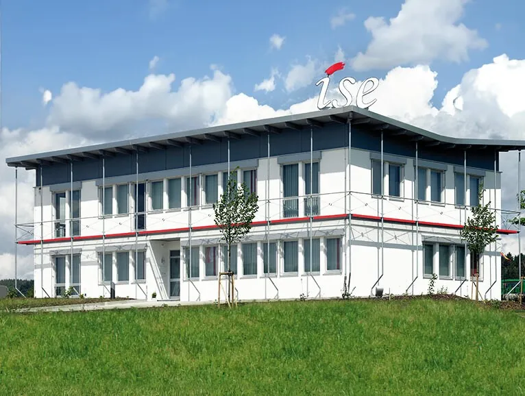 Modernes, weißes Bürogebäude mit auffälligem "ise"-Logo auf dem Dach, unter einem bewölkten blauen Himmel.