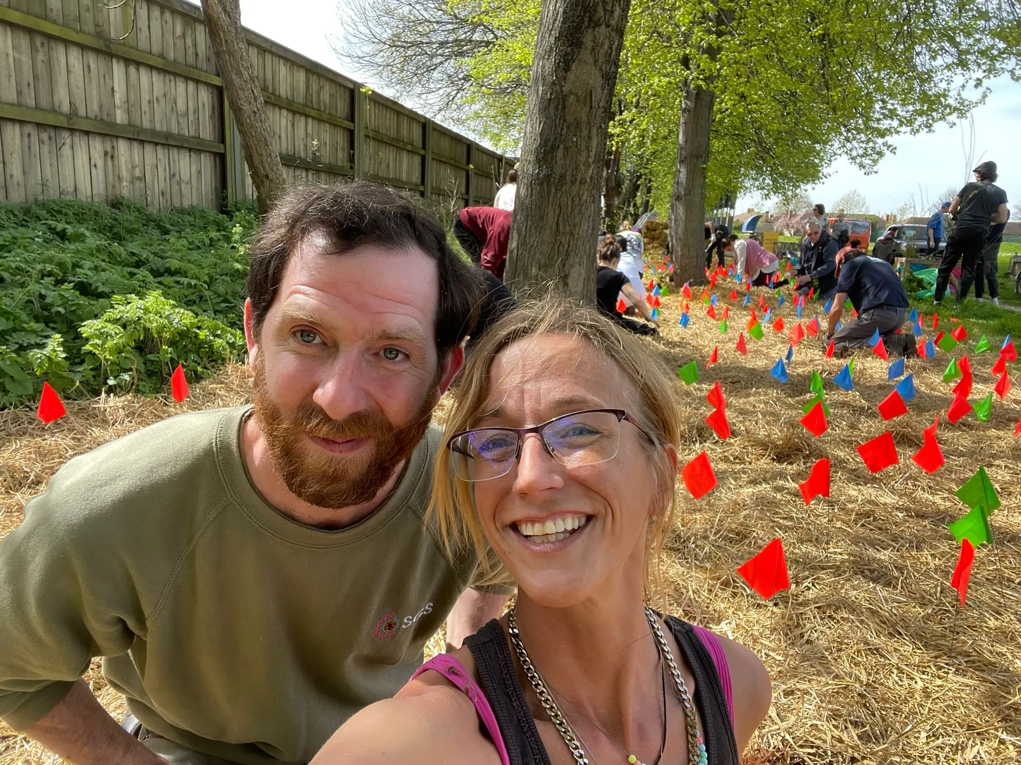 Two smiling individuals take a selfie in a vibrant outdoor setting with colorful flowers and people in the background.
