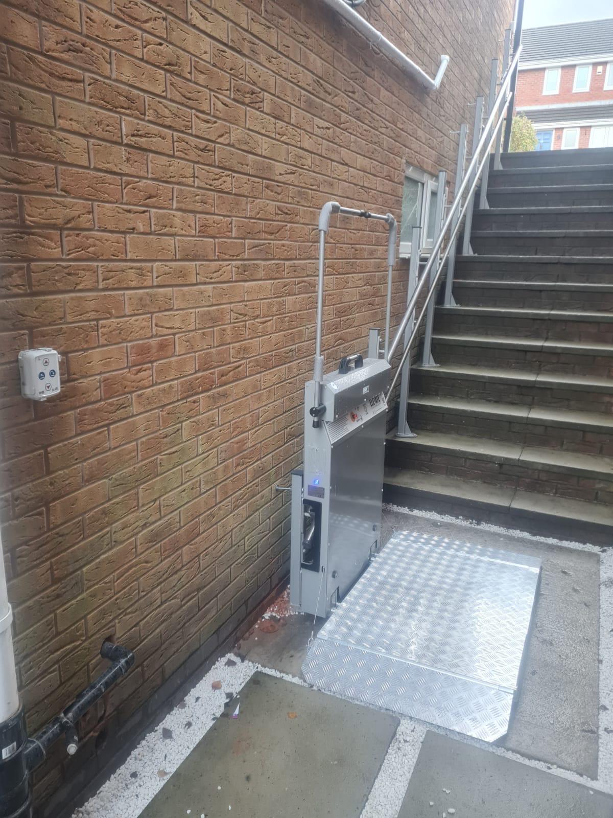 Outdoor inclined platform stairlift in raised position beside external concrete steps — wall-mounted column drive with chequerplate platform, installed at a UK commercial property with brick exterior