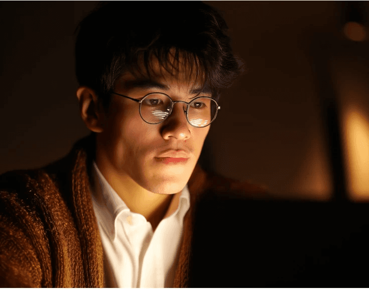 A man wearing glasses and a brown cardigan focuses intently on a laptop screen in a dimly lit room, conveying concentration and a pensive mood.