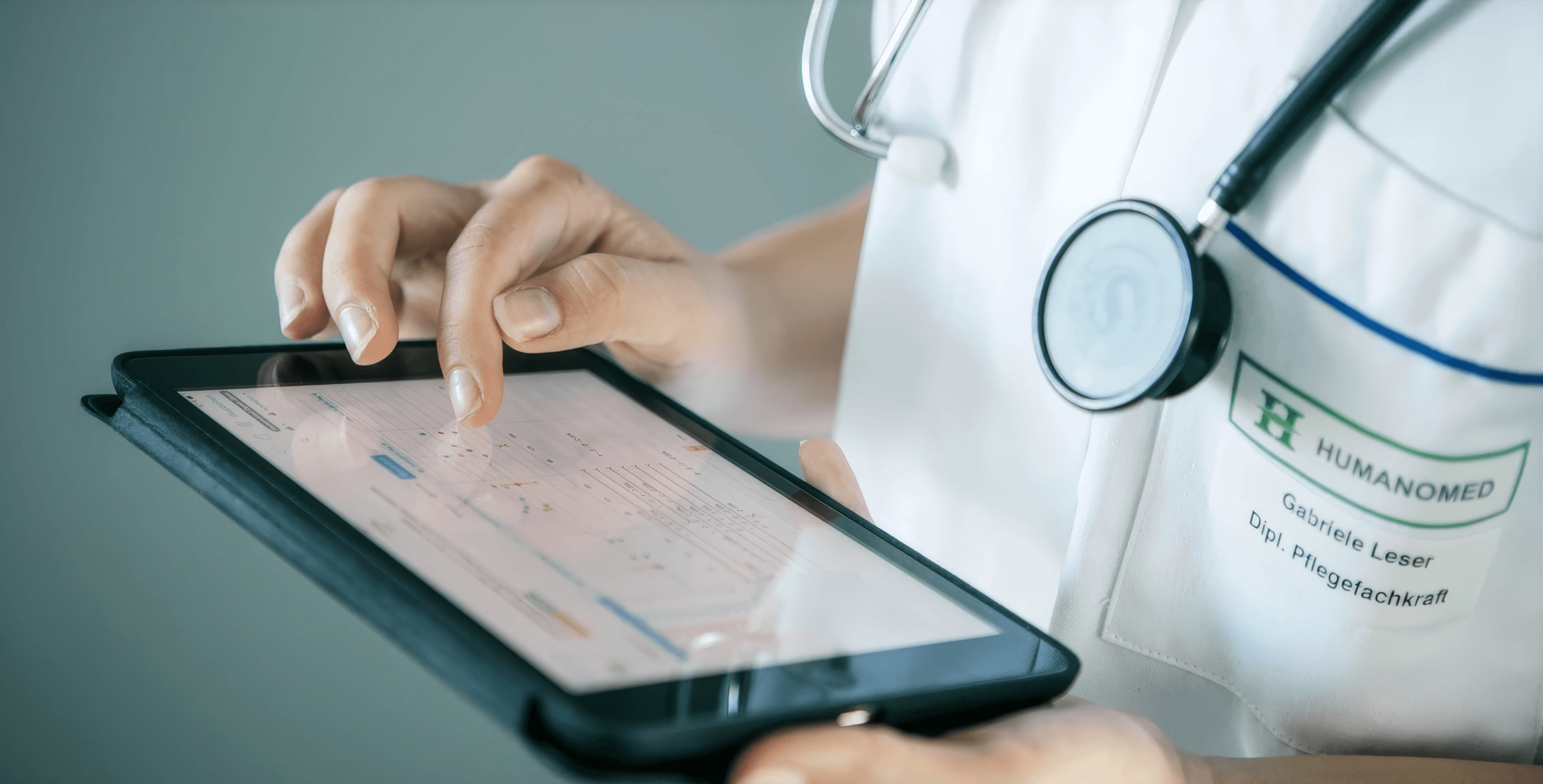 MW Interior - Pflegefachkraft mit Stethoskop und Humanomed-Namensschild bedient ein Tablet mit medizinischen Daten" Beschreibung: Humanomed-Pflegefachkraft im Einsatz mit digitalem Tablet – ein Referenzkunde aus dem Gesundheitswesen, für dessen Einrichtungen funktionale und hochwertige Objektmöblierungen realisiert wurden.