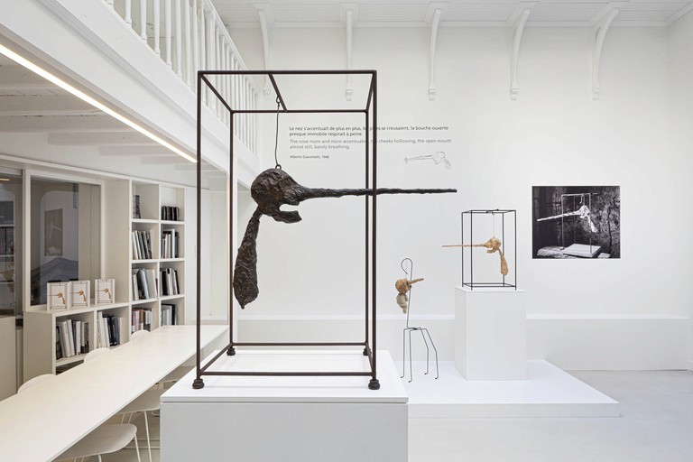 Alberto Giacometti sculptures in bright white gallery at Institut Giacometti Paris, with elongated bronze head in metal frame and archival photo on wall