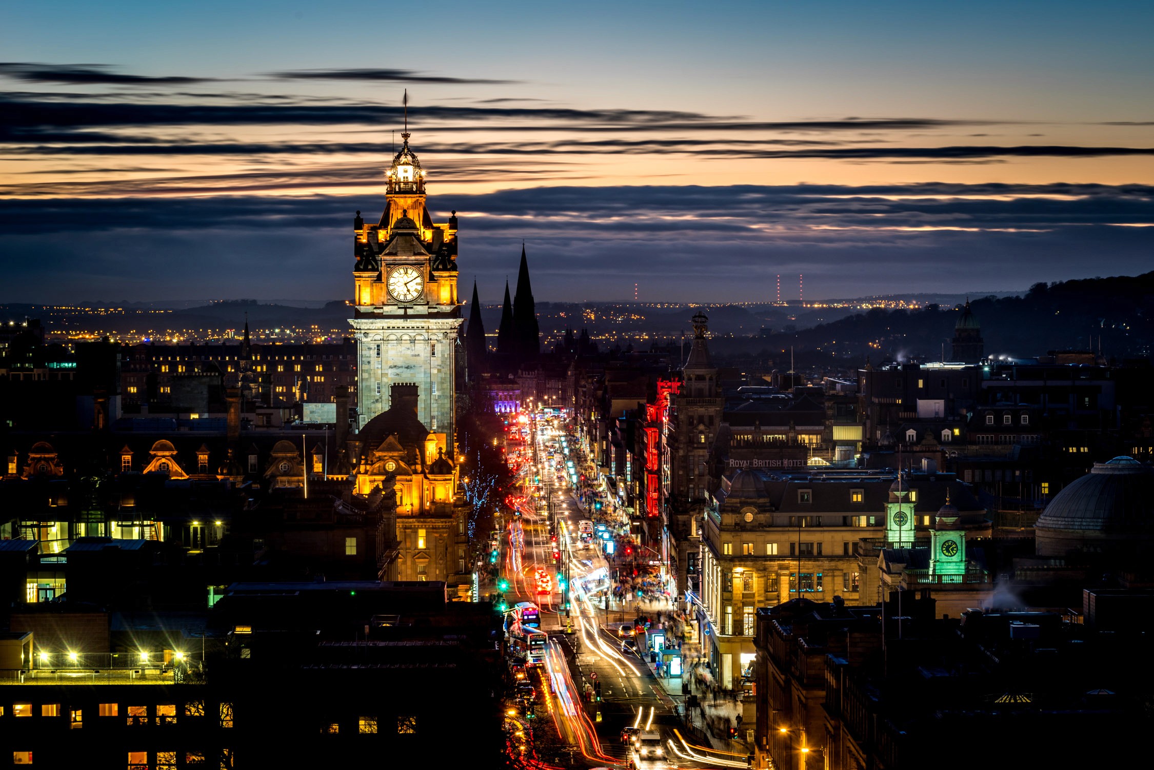 A night view of a city street with glowing lights and a clock tower under a twilight sky with horizontal clouds.