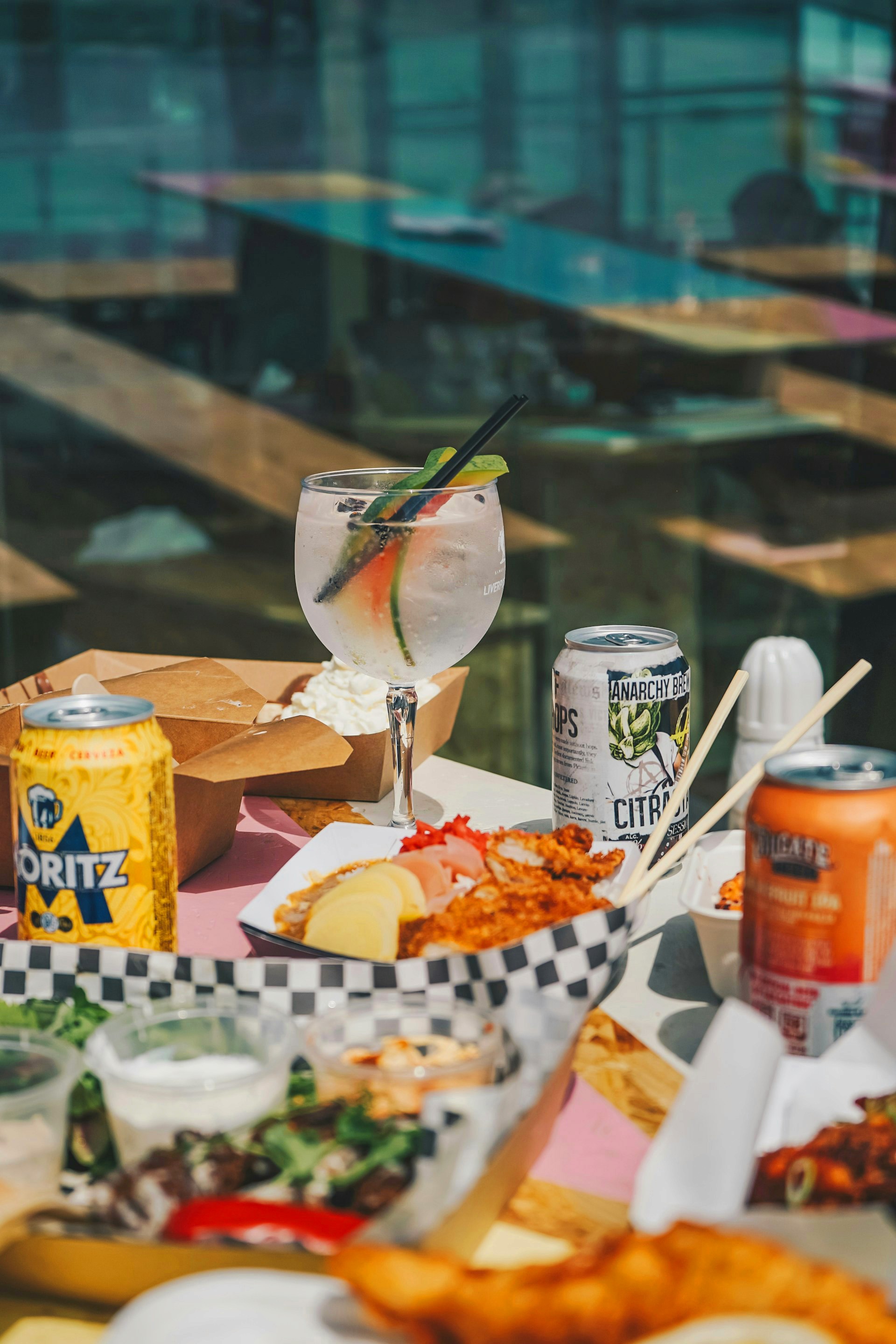 An outdoor table laden with assorted street food in cardboard containers, canned beers, and a large cocktail.