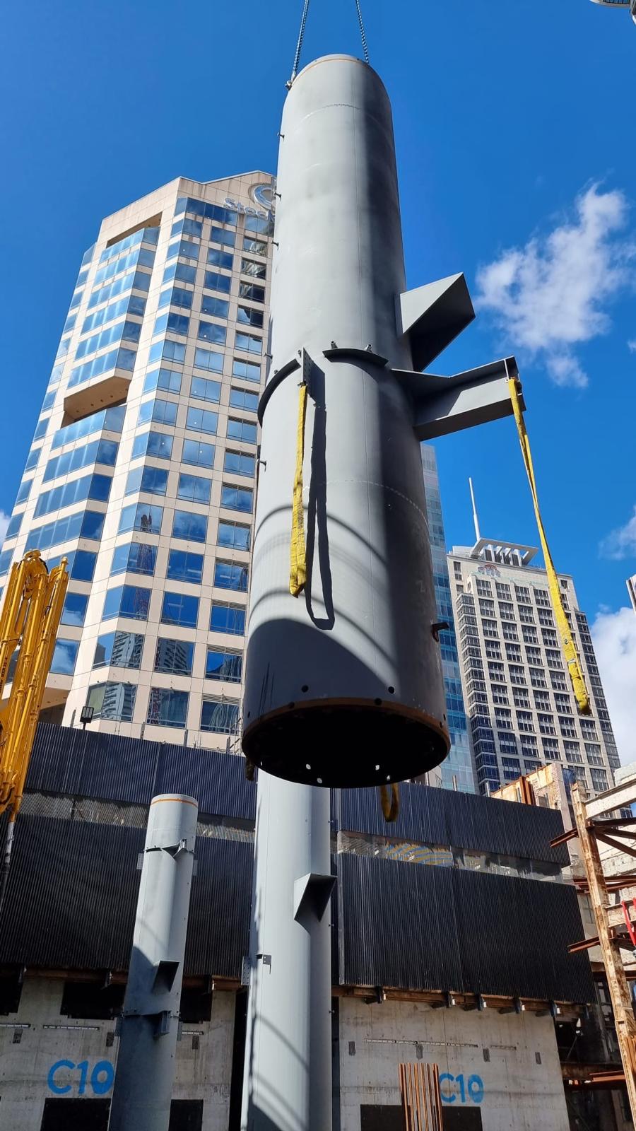 Crane lifting large metal cylinder with buildings backdrop.