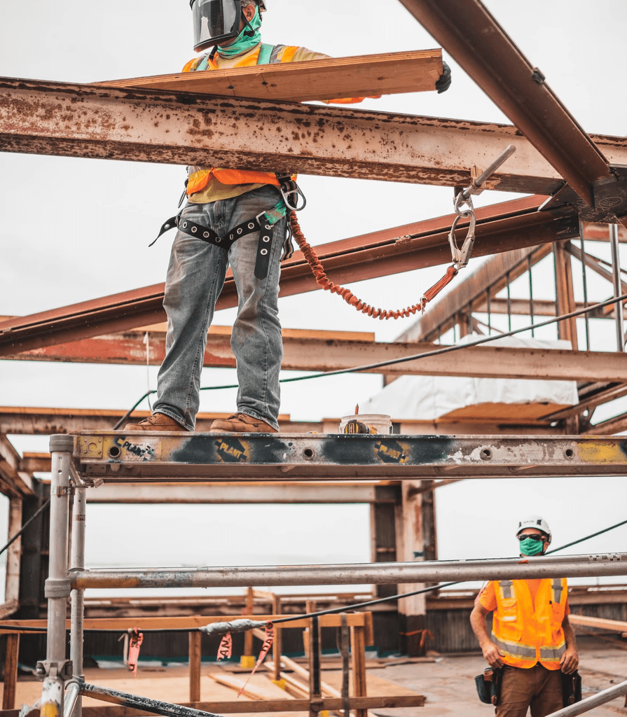Construction workers on site; one stands on scaffolding, wearing a safety harness and helmet, holding a wooden plank, conveying focus and teamwork.