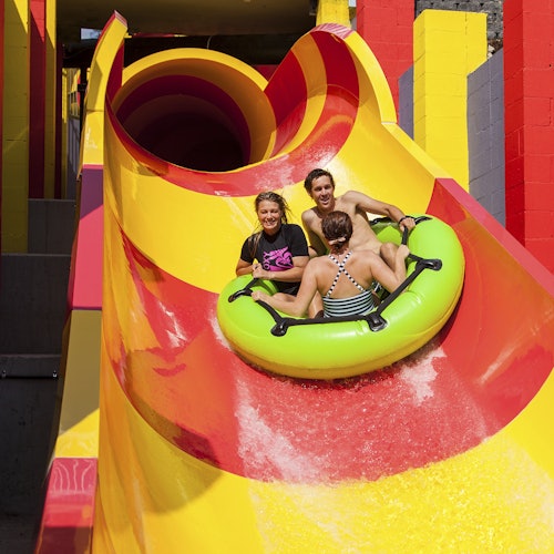 Drie personen glijden op een groene opblaasbare raft van een kleurrijke waterglijbaan in een attractiepark.