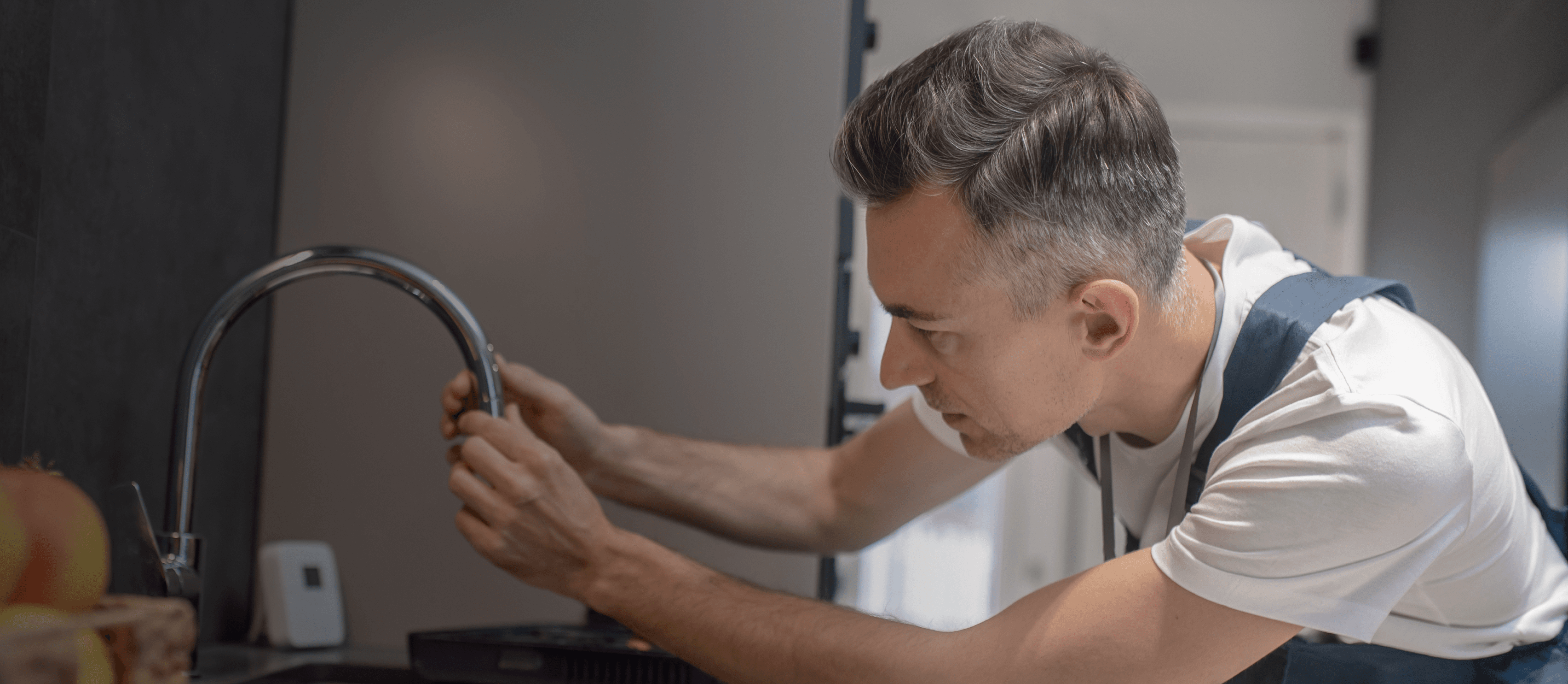 Plumber fixing a kitchen faucet. Man in white shirt and overalls adjusting plumbing. Home repair and maintenance.