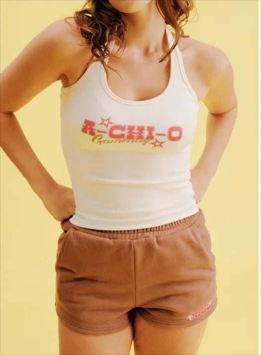 Woman in a white racerback tank top with retro-style "A-Chi-O" sorority graphic and brown shorts, standing against a soft yellow background.