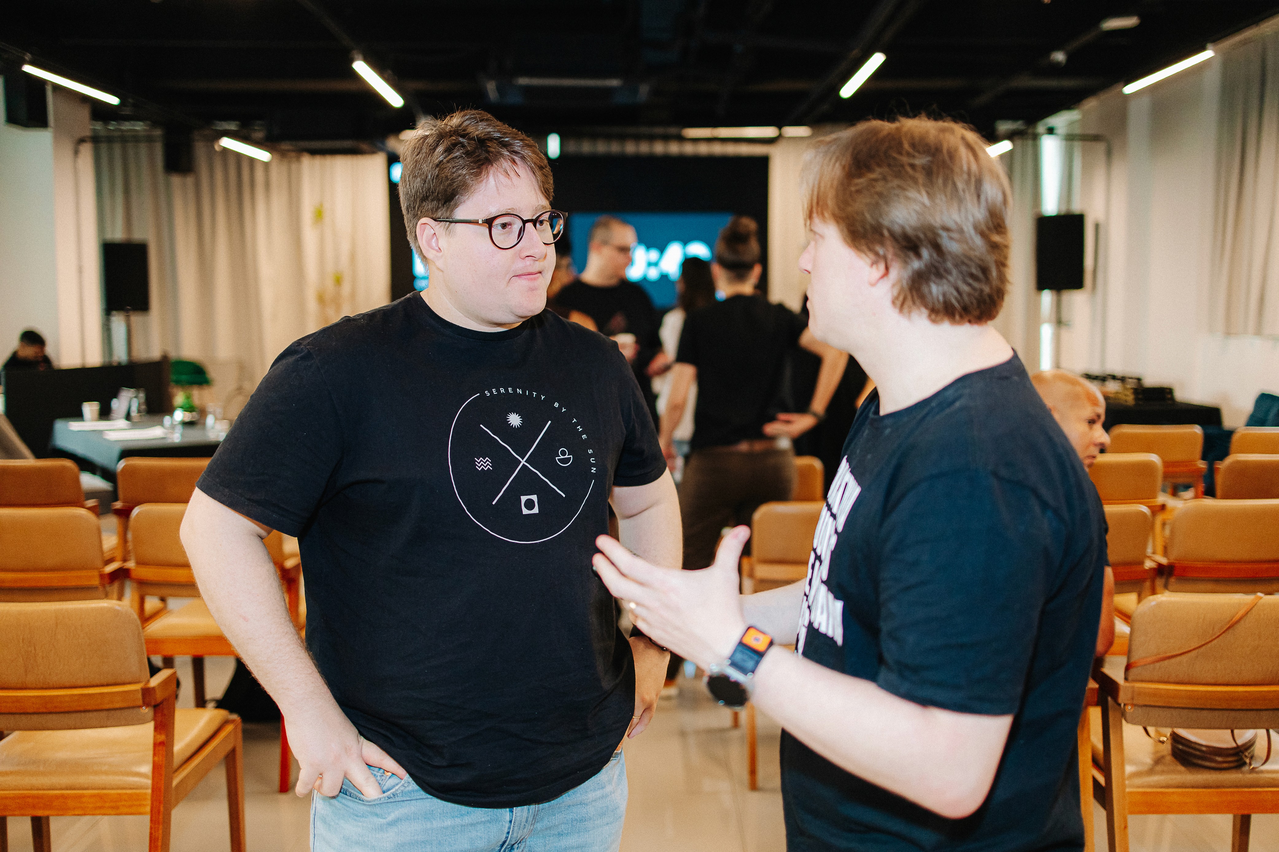 Uma pessoa está em pé no palco falando ao microfone, vestindo uma camiseta preta, com uma planta em primeiro plano.
