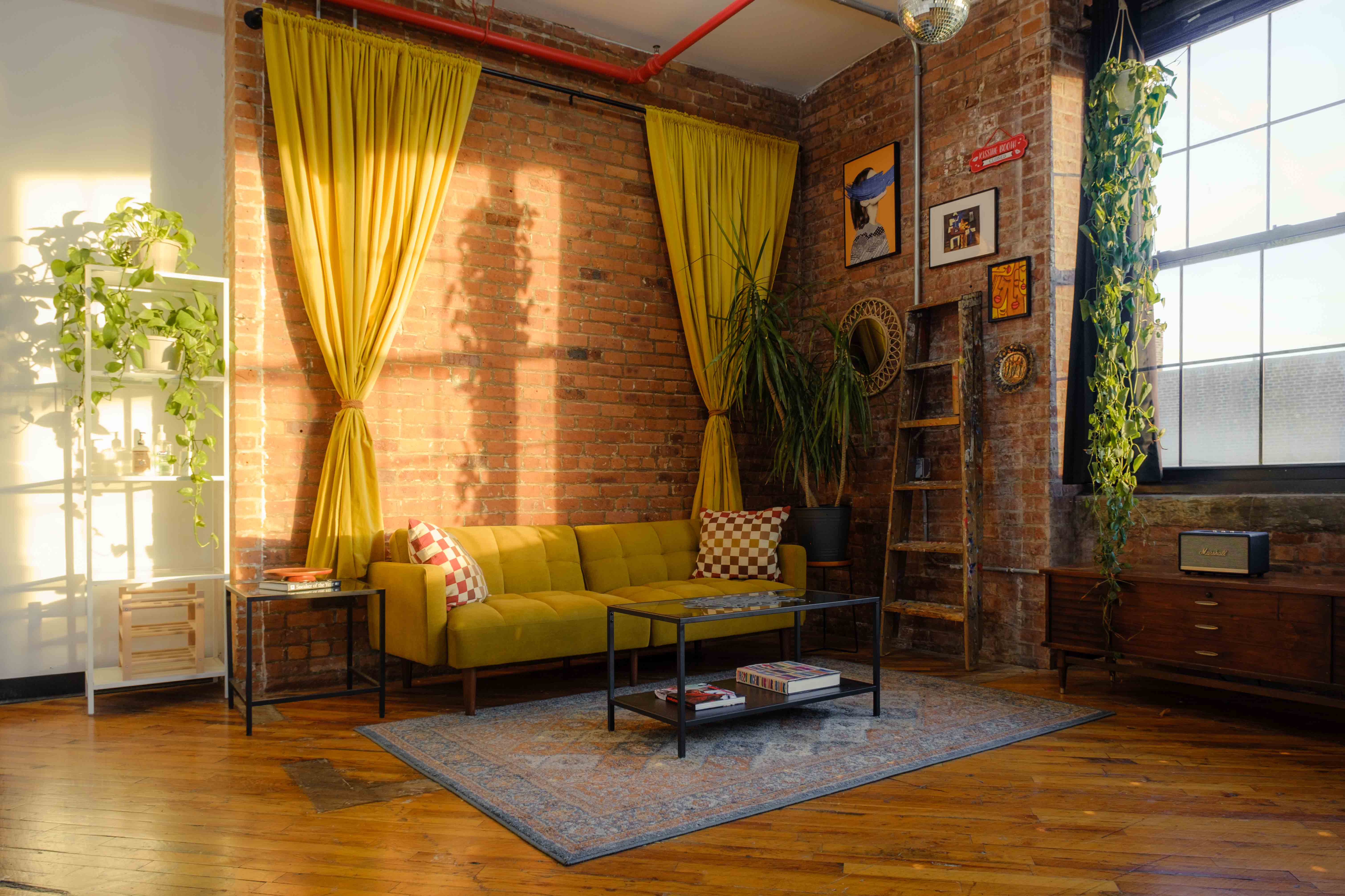 A sunlit room with exposed brick walls features a yellow couch with checkered pillows, vibrant plants, and eclectic decor, creating a cozy and inviting atmosphere. Natural light streams in from a large window with yellow curtains, adding warmth to the space.