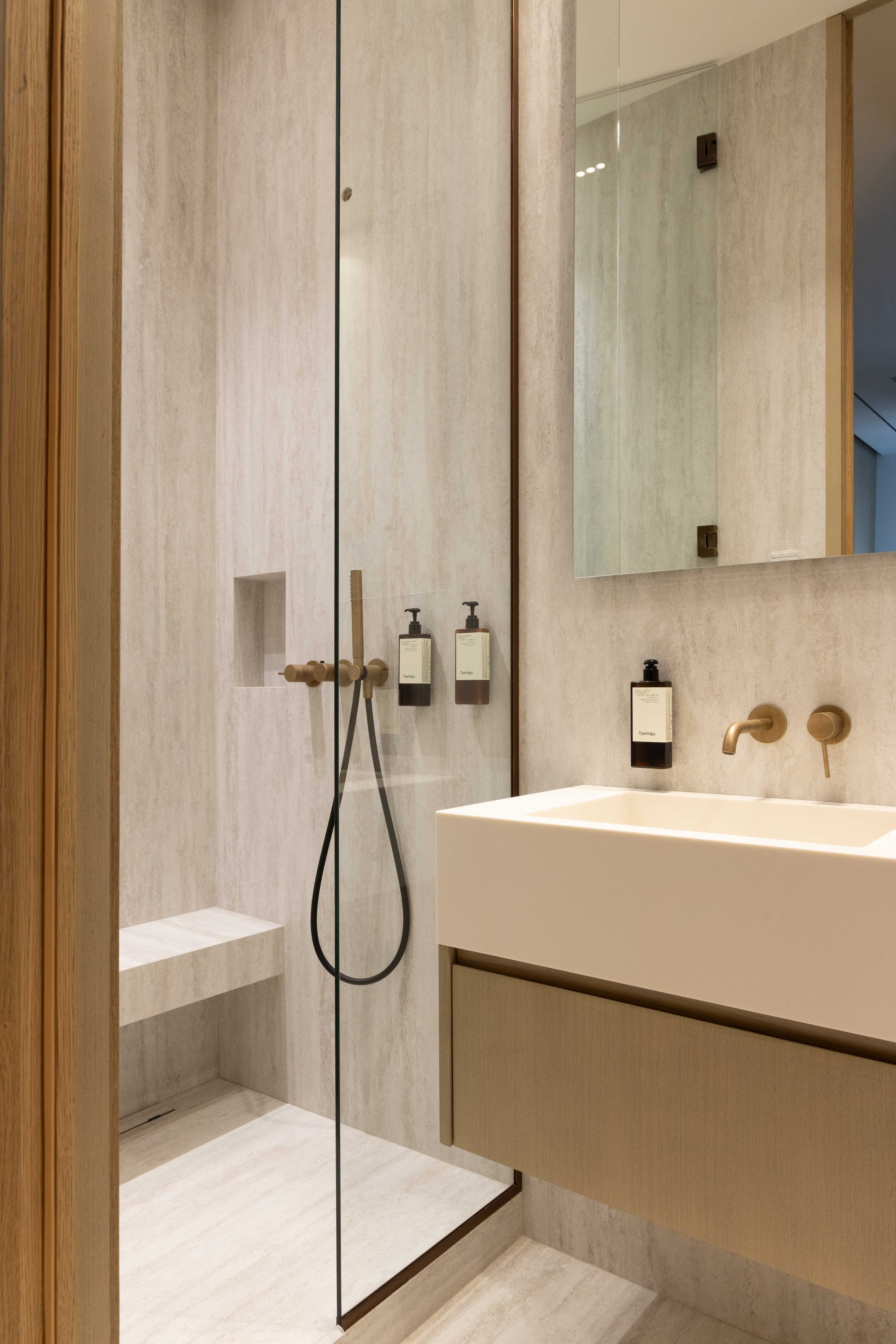 Salle de bains avec douche italienne, robinetterie doree, vasque suspendue, miroir et paroi vitree en travertin