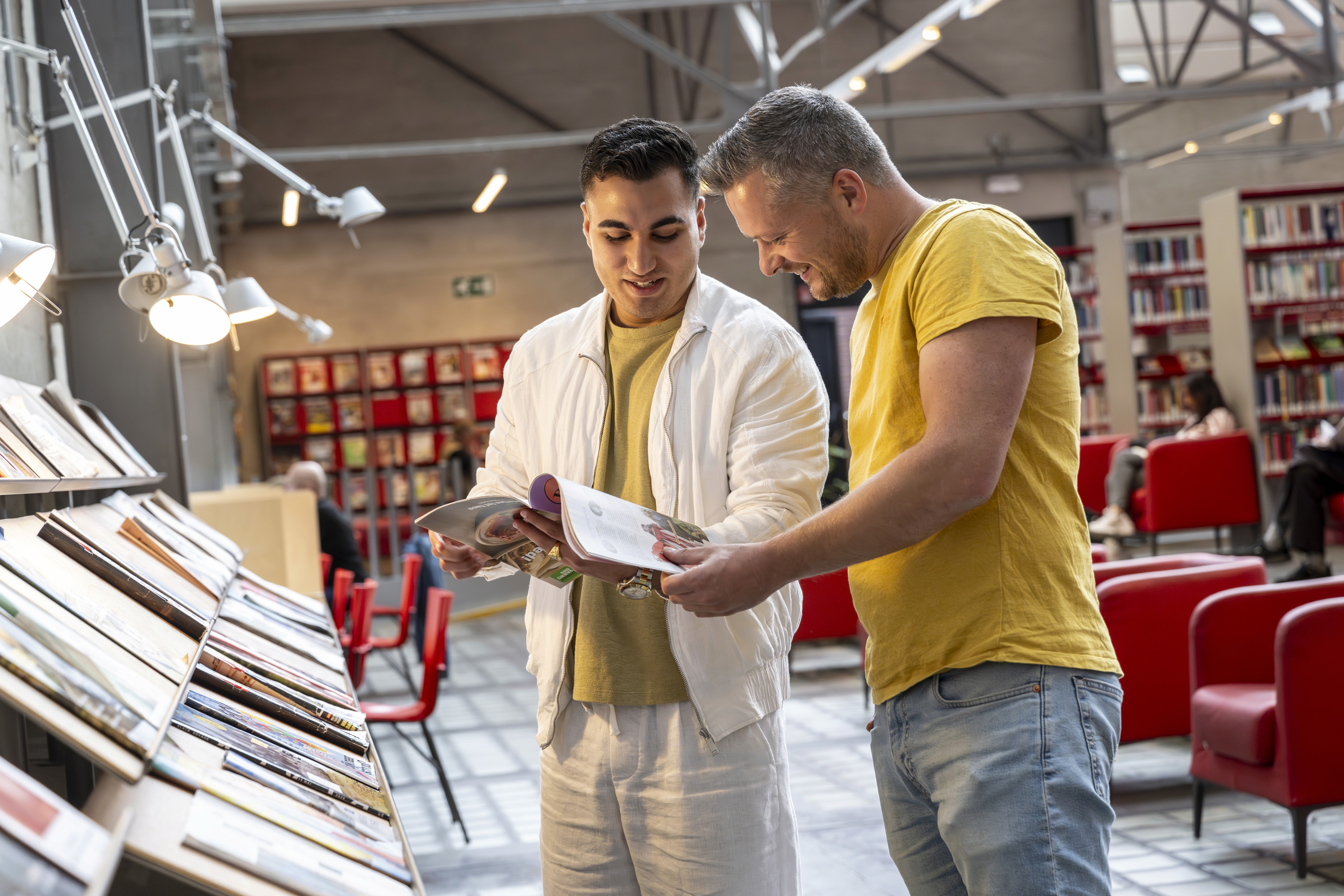 Two women on bicycles smile and chat while riding on a tree-lined path. One wears a floral dress, the other a white jacket and pants.