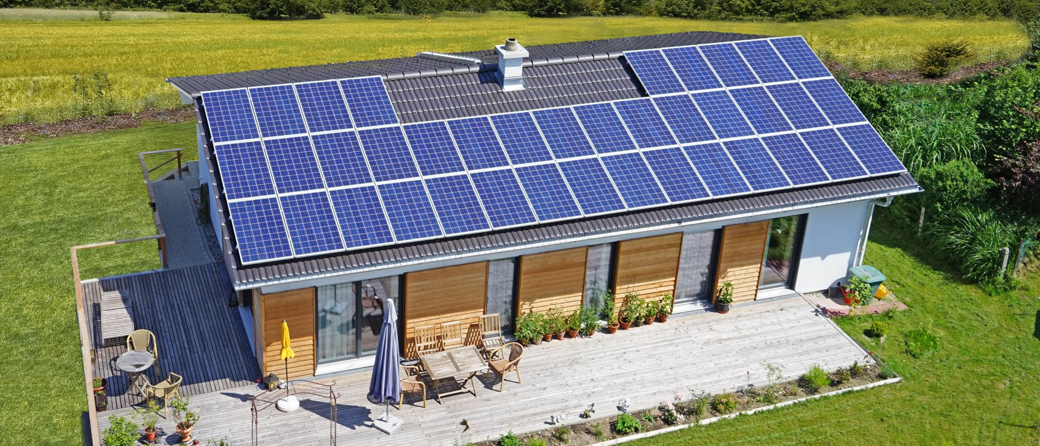 Dach mit Solarmodulen, Terrasse mit Gartenmöbeln, davor grüne Wiese und Feld.