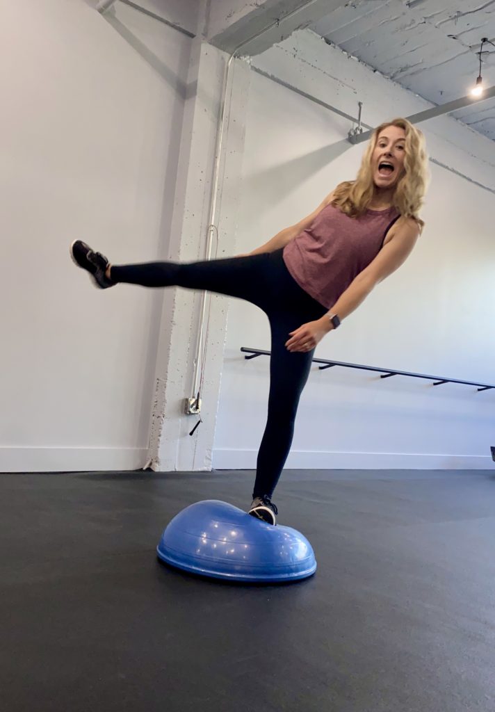 Heather Hausman with long hair is balancing on one leg on a blue balance trainer inside a brightly lit gym, wearing black leggings and a sleeveless maroon top, with a ballet barre visible in the background.