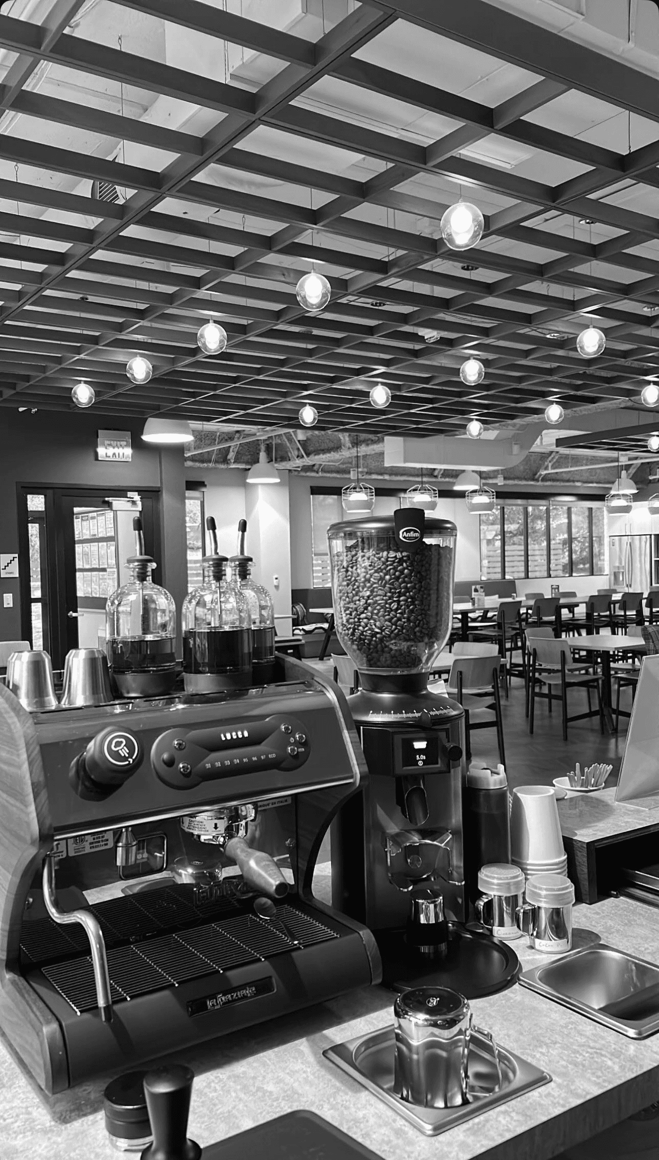 Black and white photo of a man and woman standing on stone blocks overlooking a cityscape and river. The man, dressed in a striped button-down shirt and light-colored pants, stands on the left. The woman, in an off-the-shoulder white dress, stands on the right with one hand on her shoulder. Behind them, a scenic view of a European city with buildings on hills and a river winding through the center is visible at sunset or twilight. A railing lines the overlook where they are standing.