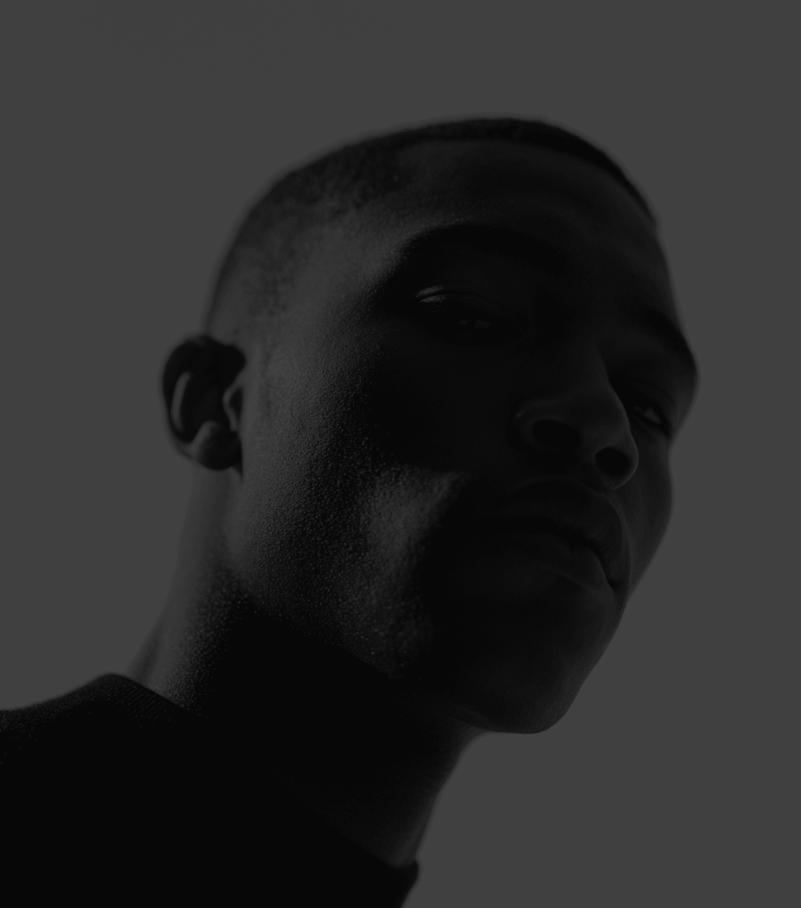 Low-key portrait of a man's face cast in deep shadow against a dark gray background.