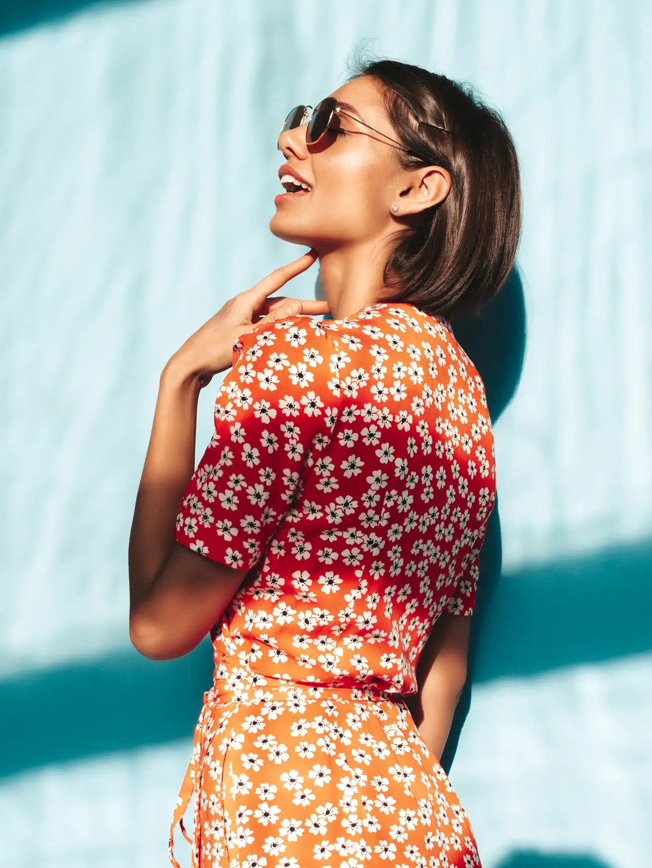 Woman in a red daisy-print summer dress laughing against a sunlit pale blue wall