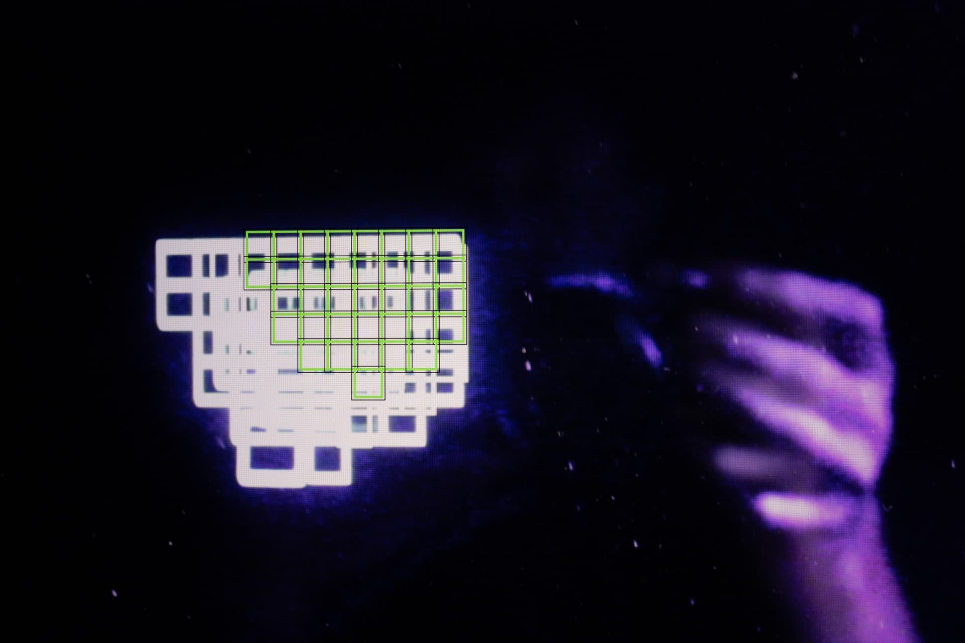 Green autofocus grid attempting to lock onto white interface element on screen, purple hand gesture visible at right creating recursive feedback between camera and display
