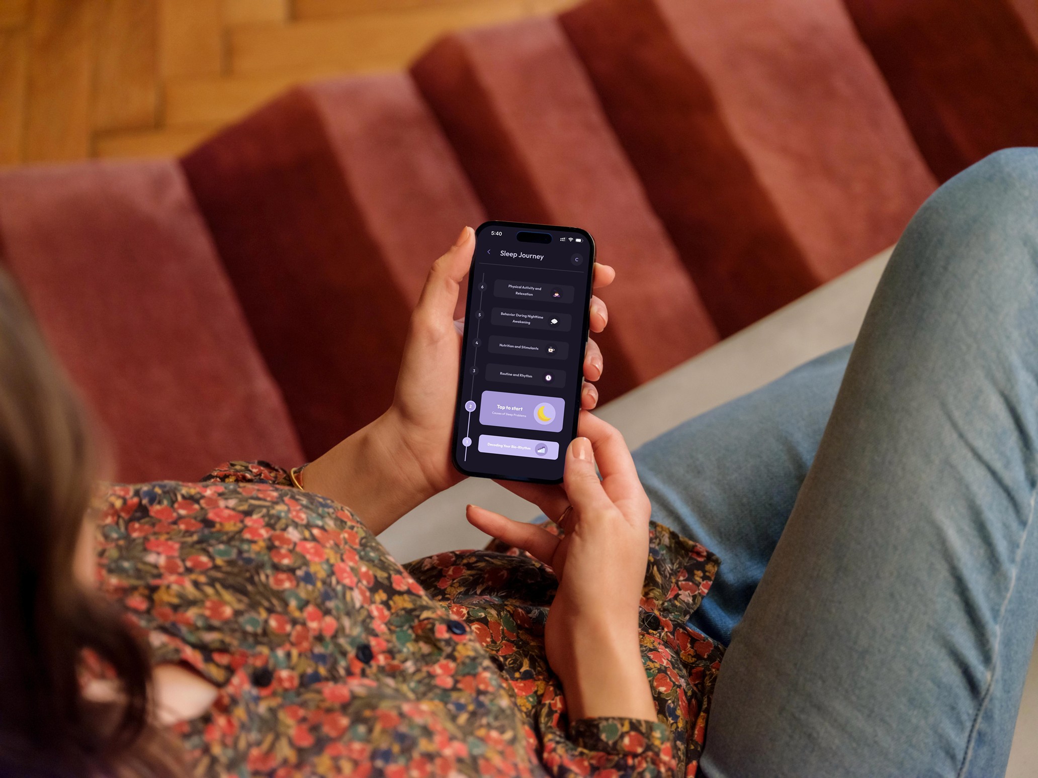 Over-the-shoulder view of a woman holding a smartphone with both hands. She is wearing a floral blouse and blue jeans, sitting in a cozy environment with red upholstery. The phone screen clearly displays the Vialog app's "Sleep Journey" timeline with the yellow moon icon at the bottom.