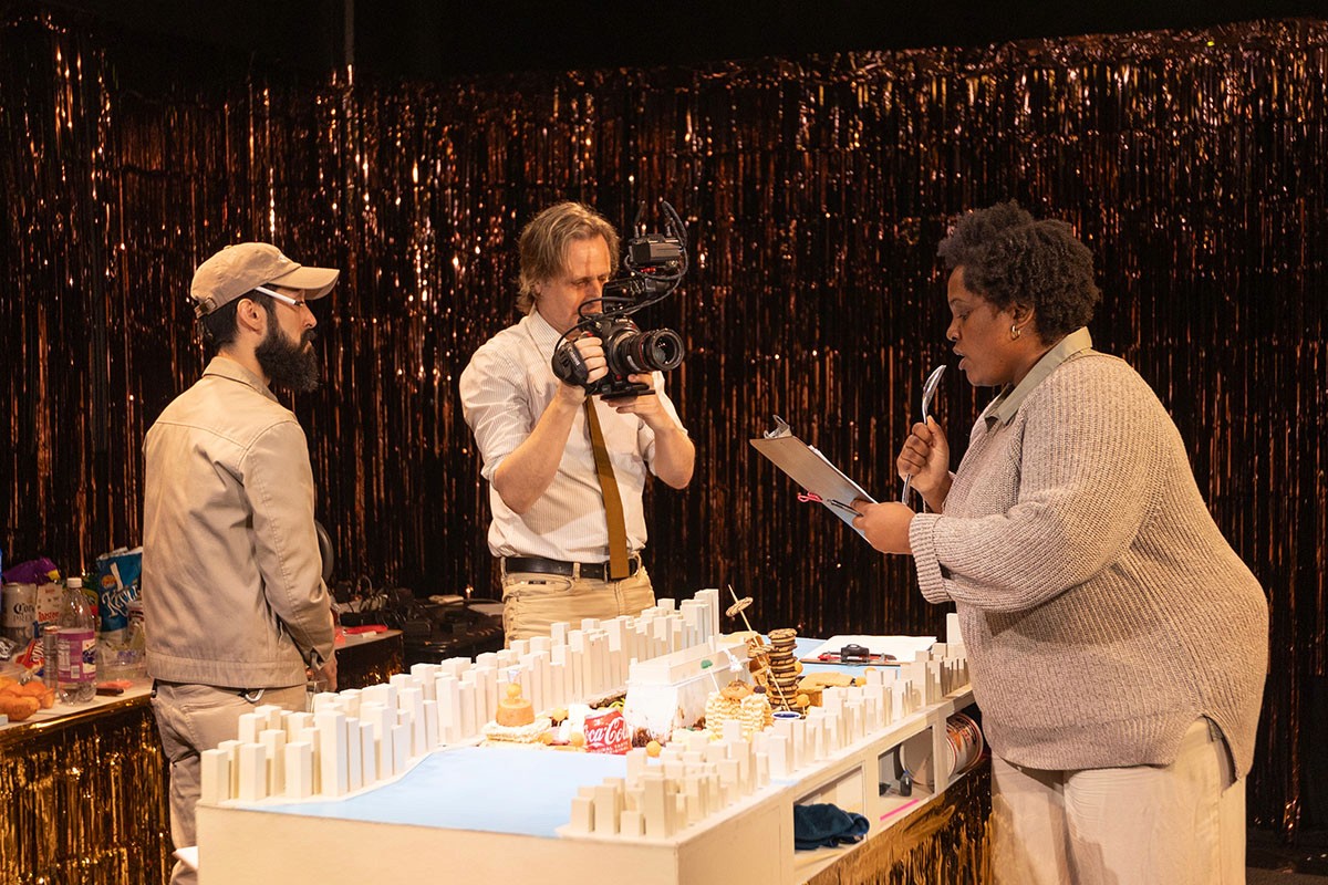 Three performers act out an interview scene. The center performer holds a camera.