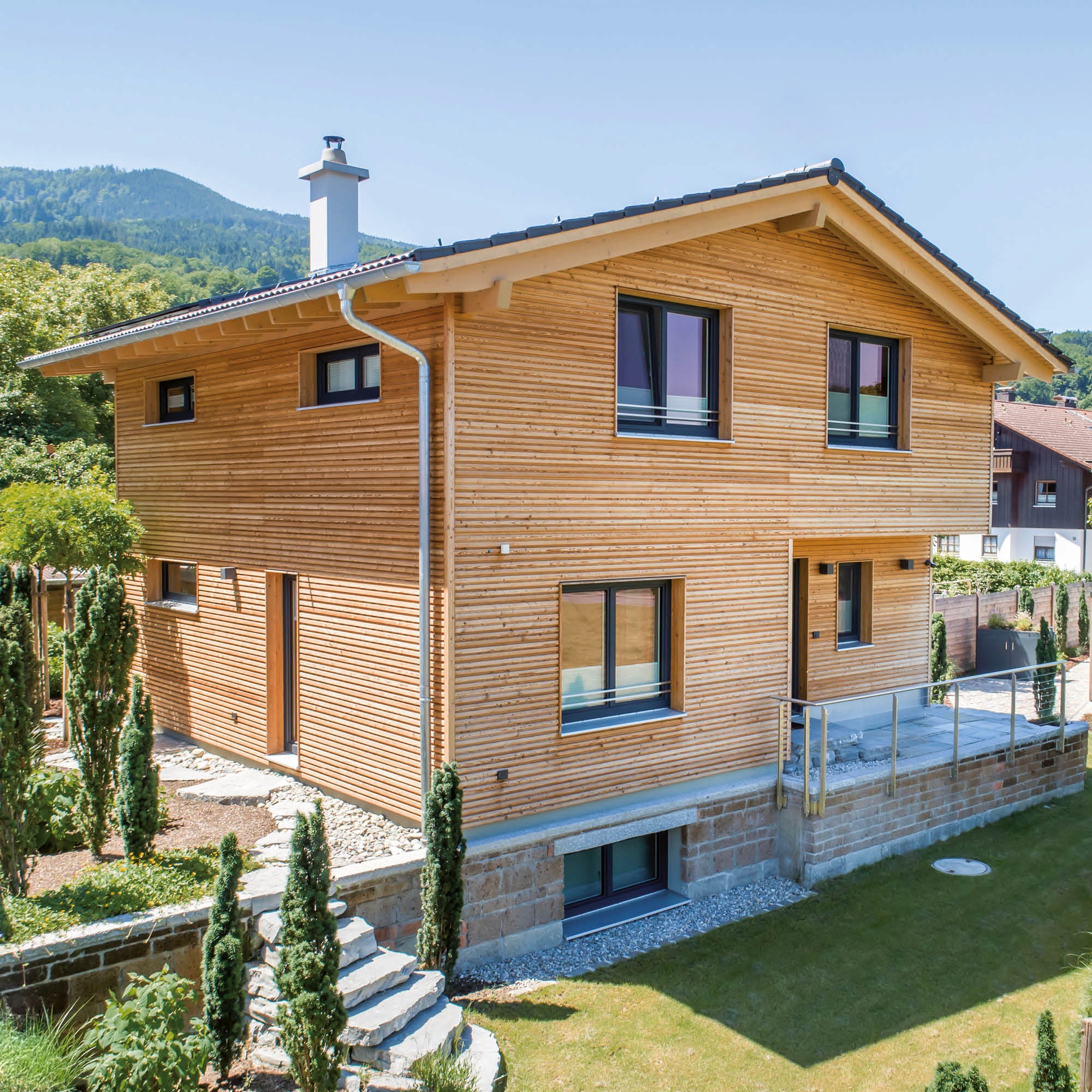 Modernes Holzhaus mit anthrazitfarbenen Fenstern und einem Mansarddach, umgeben von Grünflächen und Bäumen.