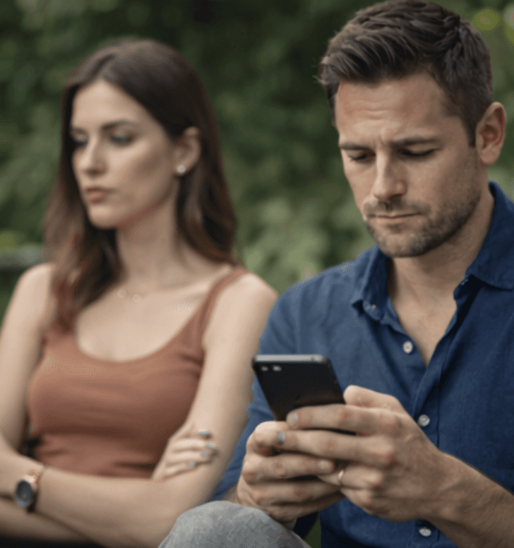 ein mann schreibt an seinem handy, eine genervte frau sitzt neben ihm