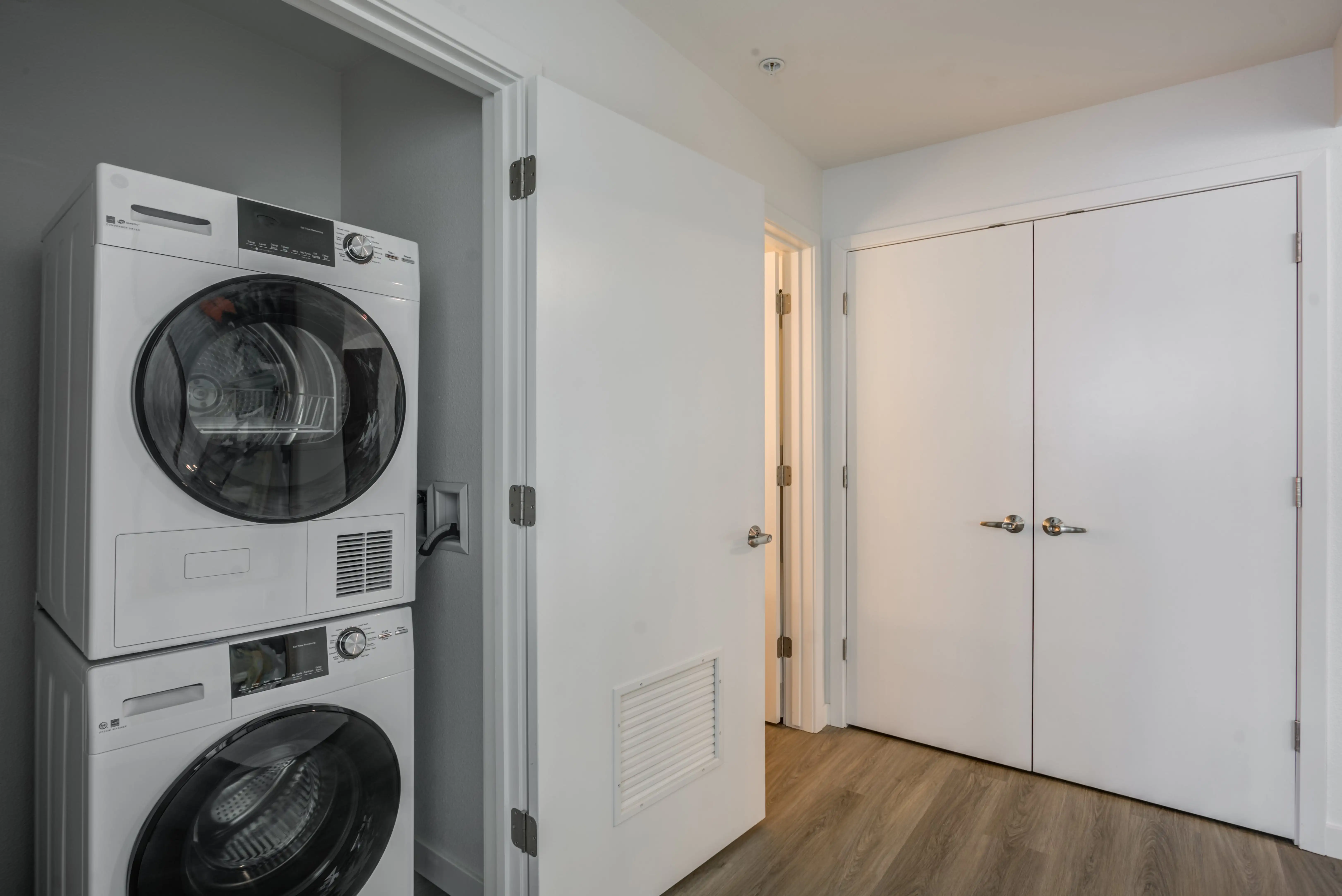 A laundry area in a hallway features a stacked washer and dryer unit next to a partly open door. Adjacent to this door is a closed double-door closet. The sun-drenched space is well-lit, with wooden flooring and white walls, perfect for the modern living spaces found in Midtown Sacramento.