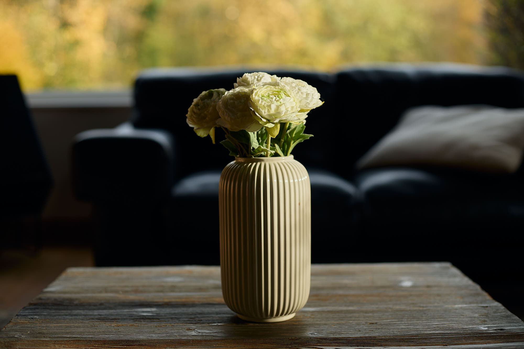 Gros plan d'un vase blanc avec des lignes verticales style art déco, avec des fausses fleurs blanches à l'intérieur, posées sur une table basse en bois avec en arrière-plan flou, le canapé noir en cuir du salon, devant la baie-vitrée.